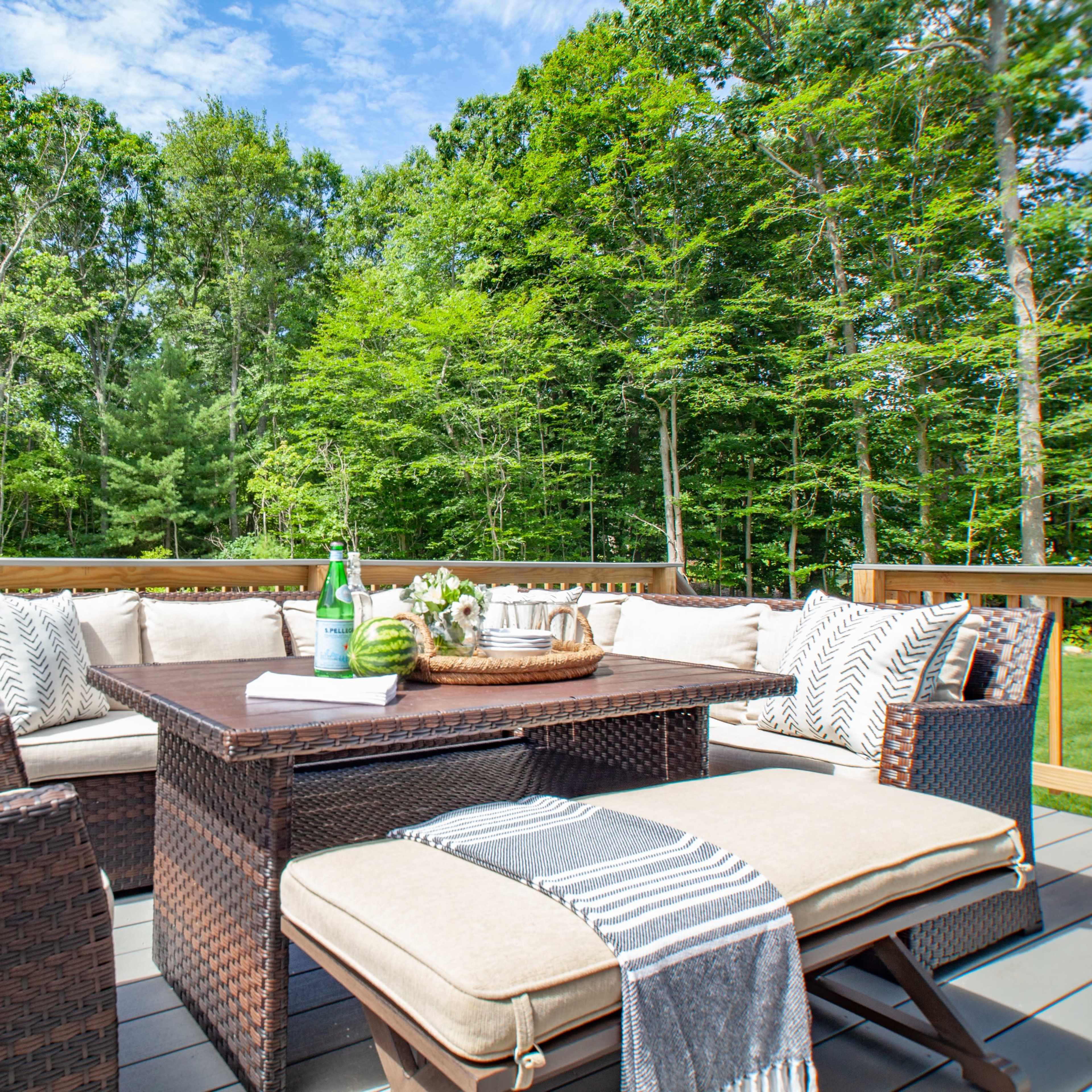 A furnished outdoor deck features a woven seating arrangement with cushions, a central coffee table, and potted plants surrounded by trees.