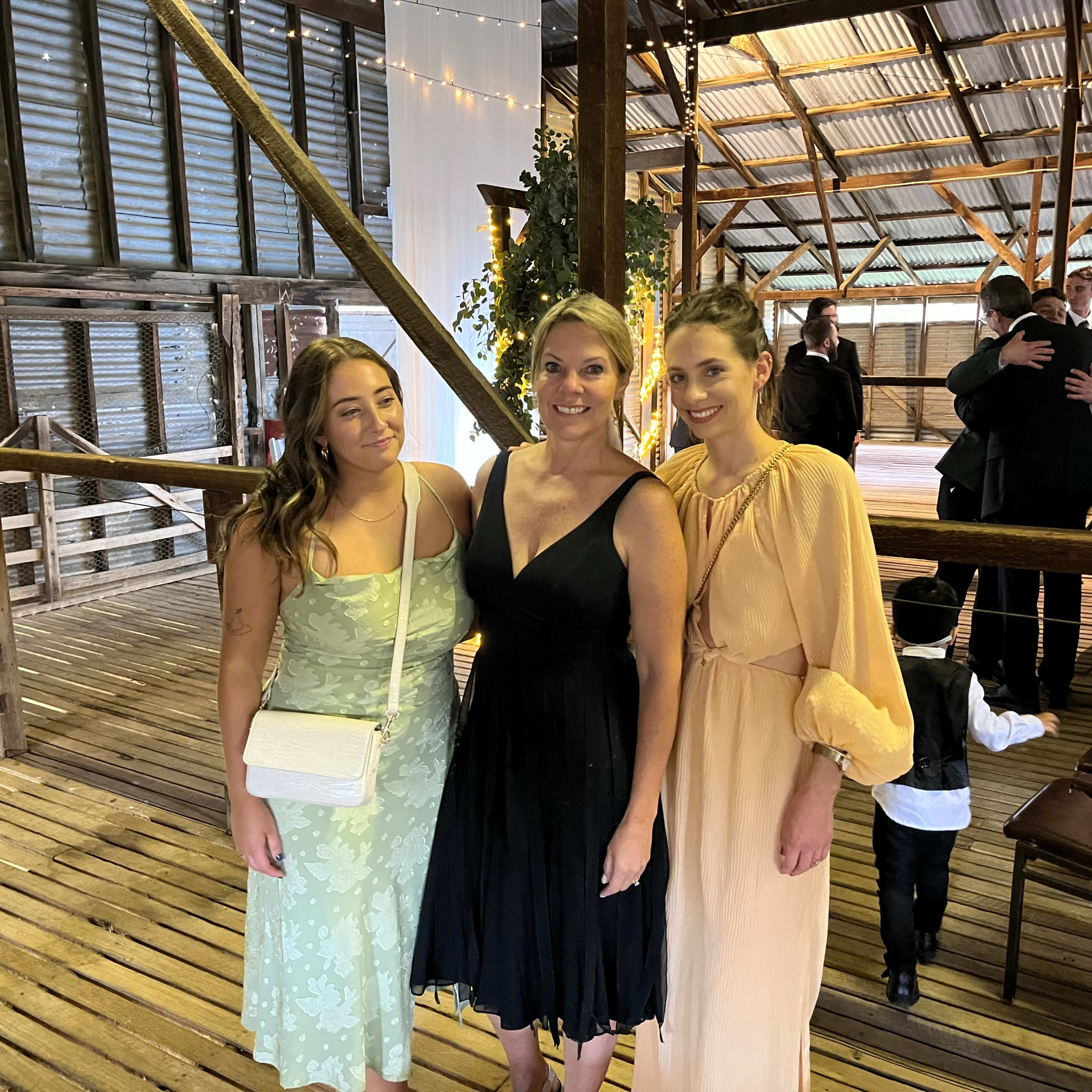 Three women pose together in a rustic barn setting at a social event.