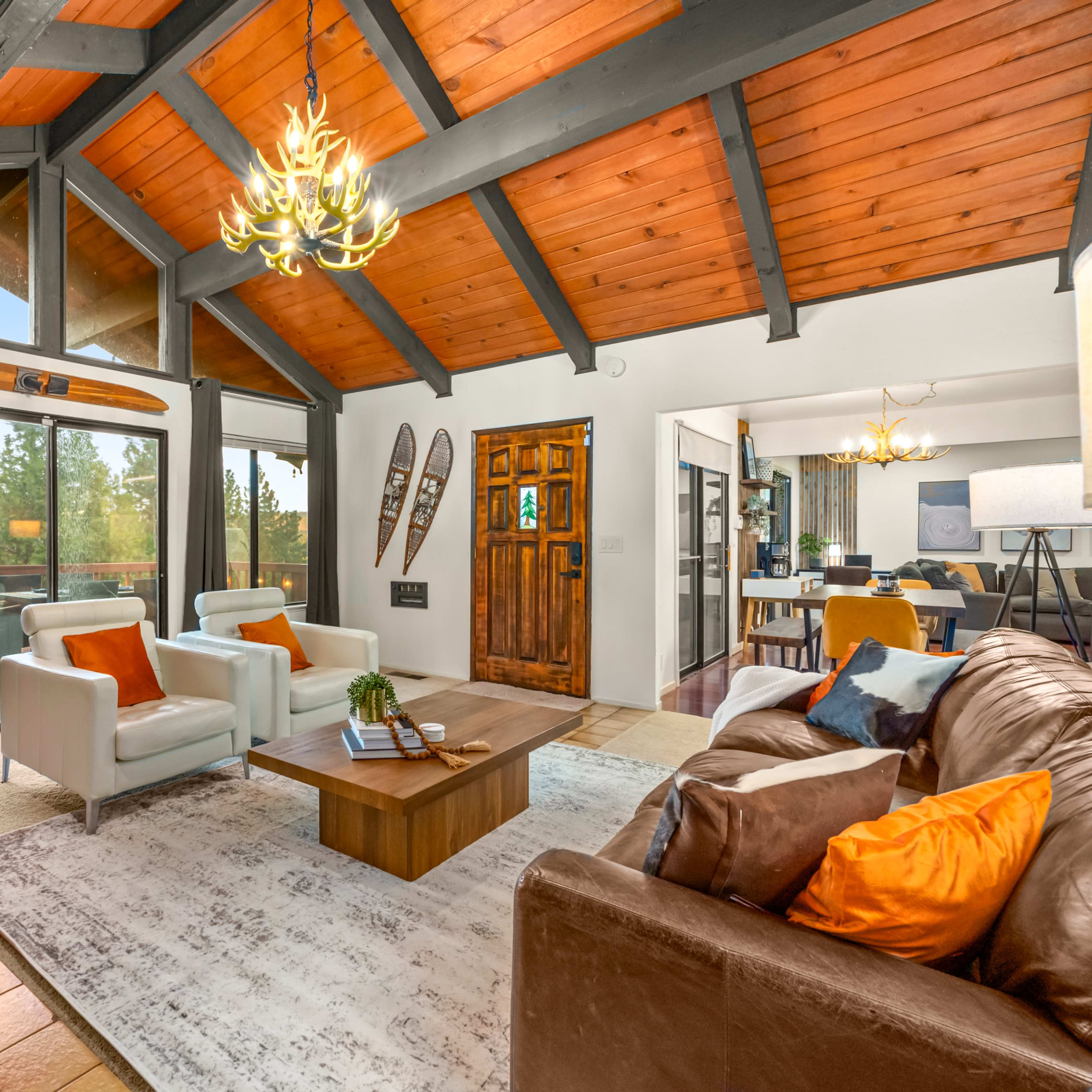 The living room features a high-beamed ceiling, large windows with a view of the outdoors, a central wooden coffee table, and a mix of seating arrangements.