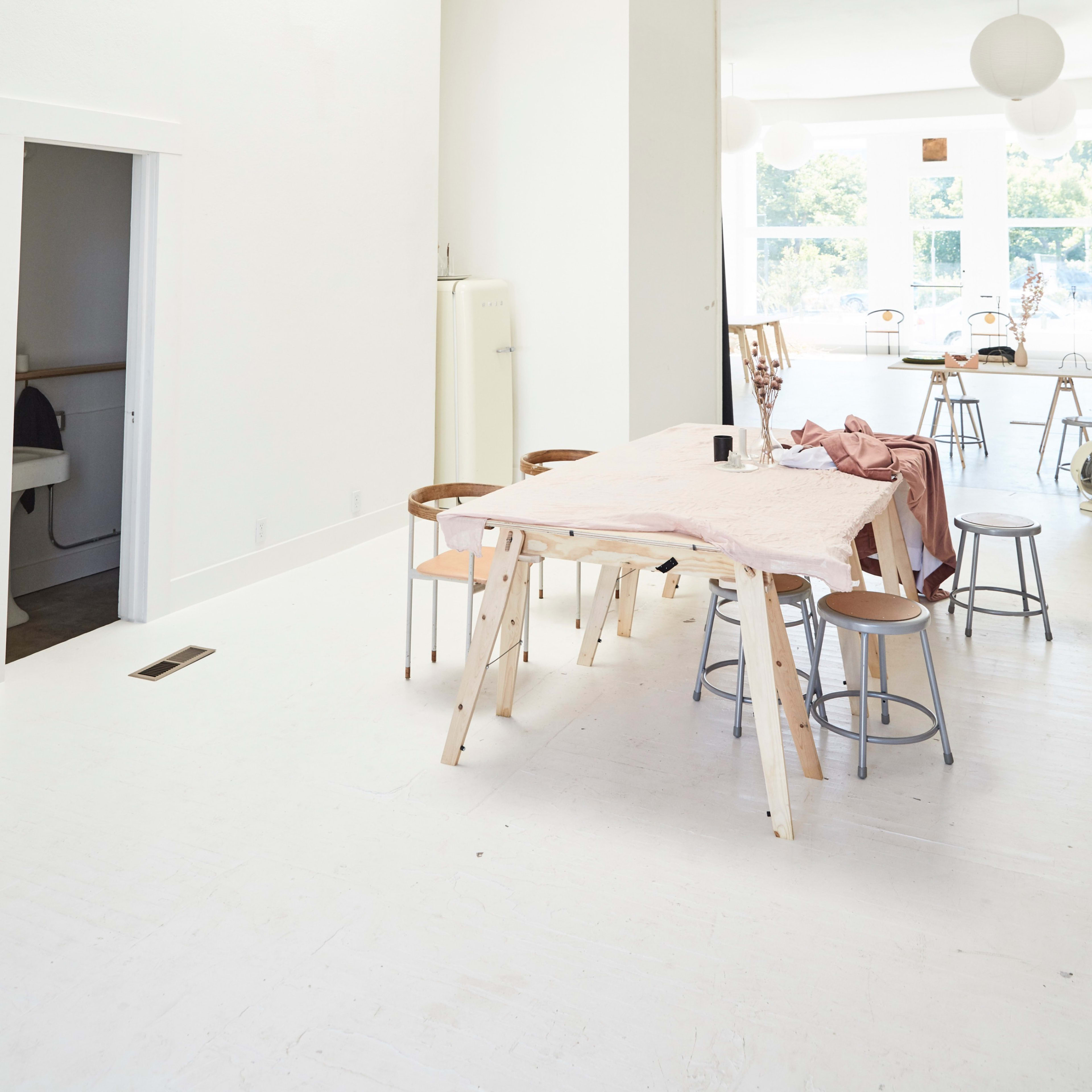 A bright, minimalist space features a central wooden table surrounded by chairs, with large windows allowing natural light to fill the room.