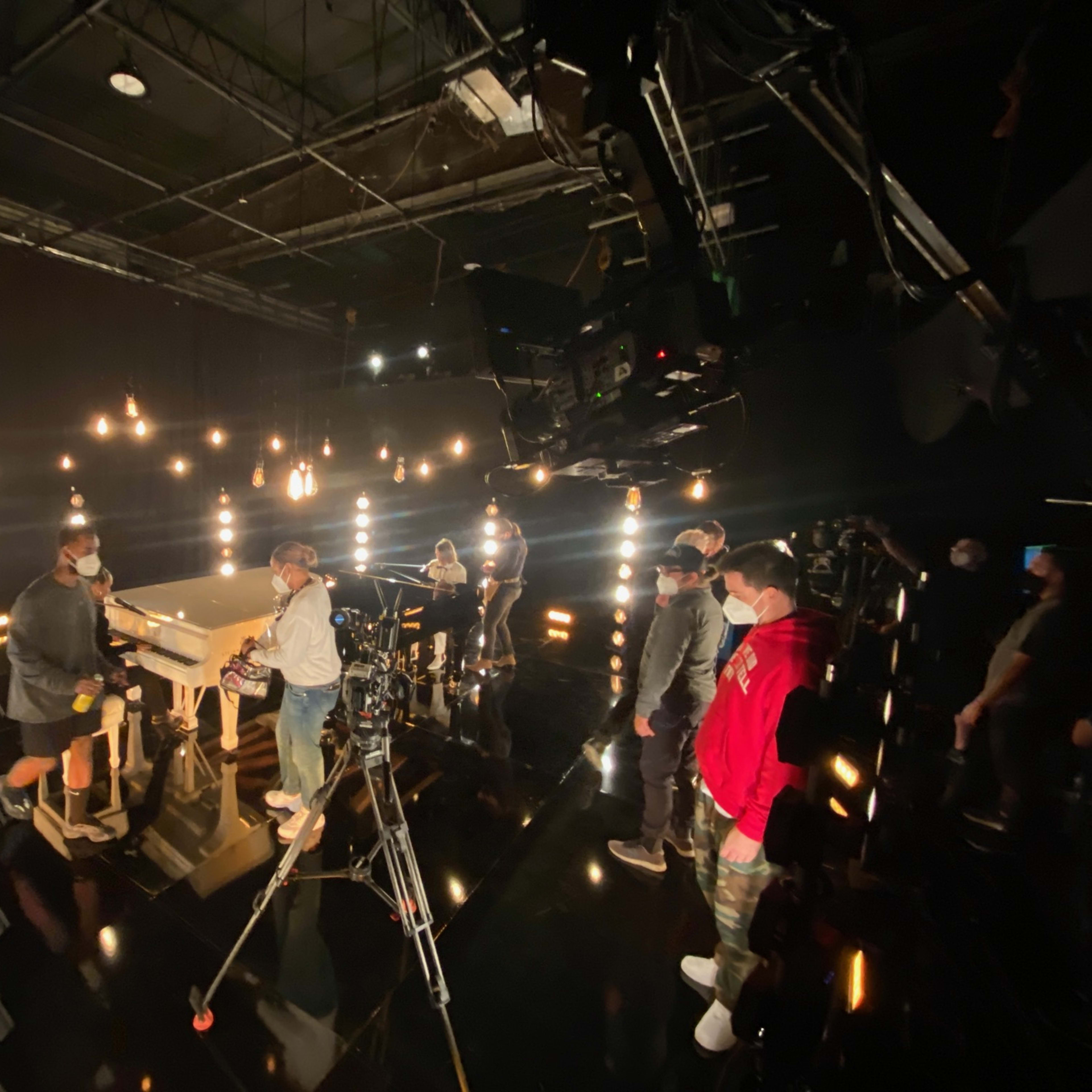A film crew works around a central table in a dimly lit studio, with lights suspended above and cameras positioned for a shoot.