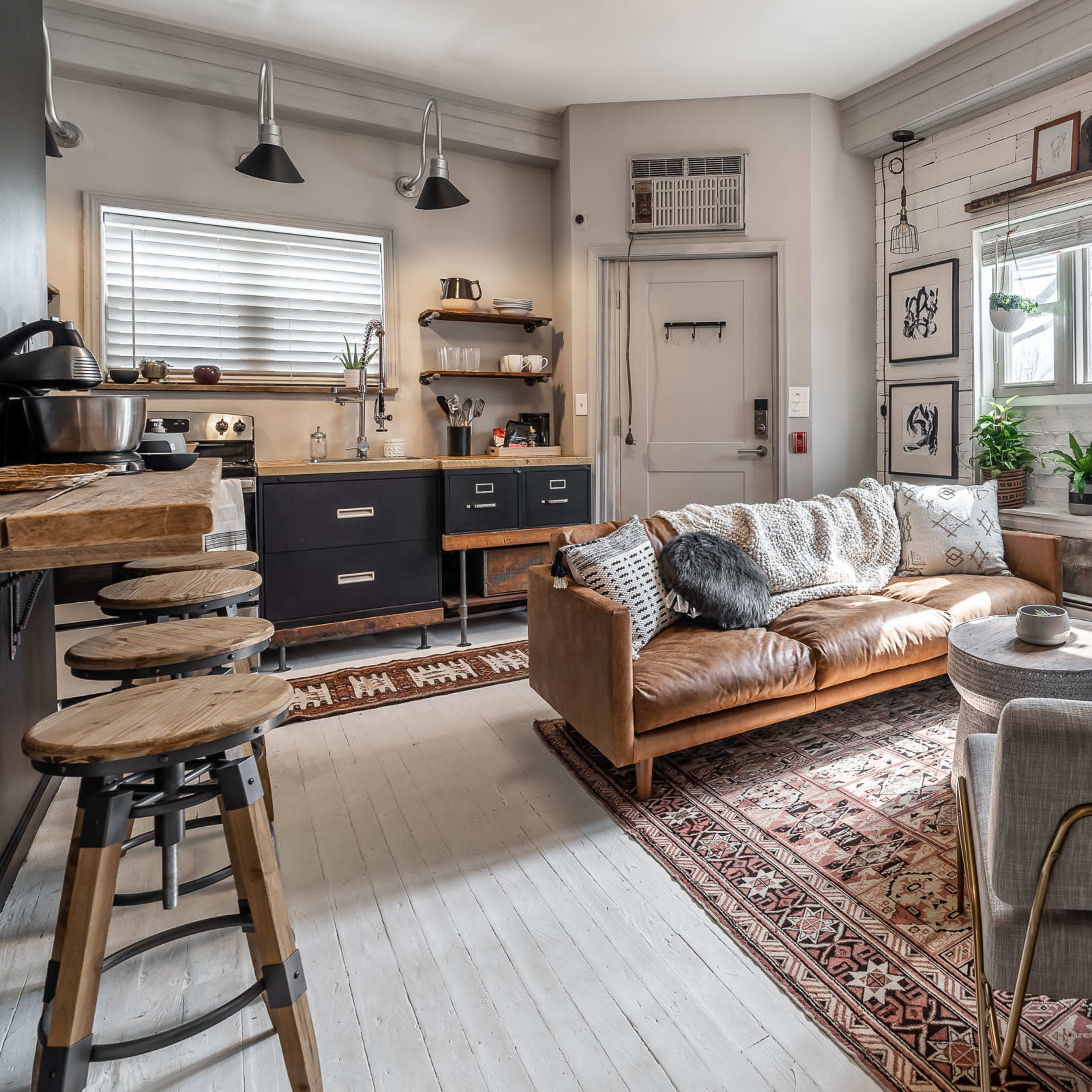 A cozy living room features a leather couch, a gray armchair, and a decorative rug, with framed art on the walls and a window allowing natural light to enter.