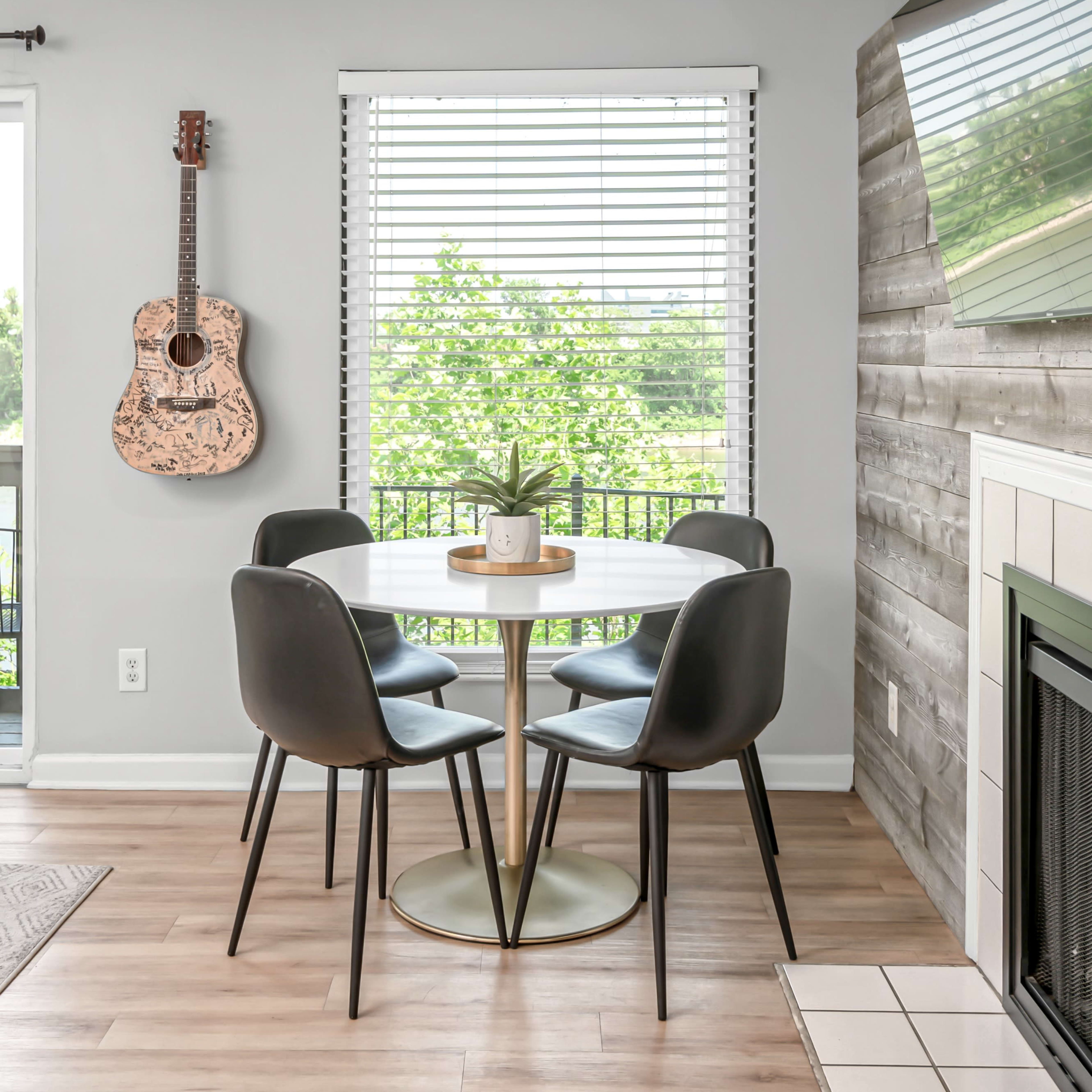 A circular dining table with four black chairs is positioned near a window with blinds, overlooking greenery, and a guitar hangs on the wall beside a fireplace.