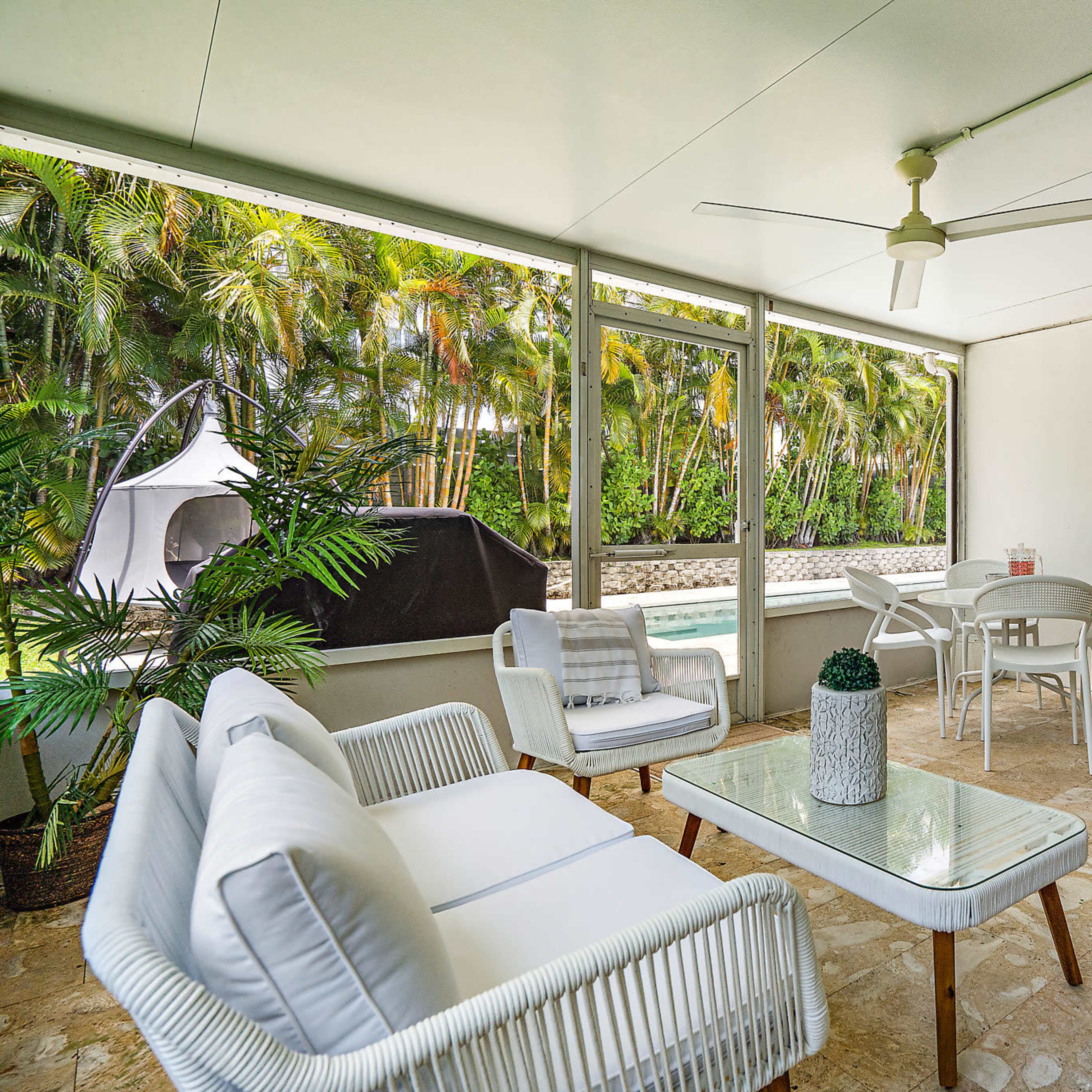 A screened patio features white wicker furniture, a glass table, and a view of a landscaped area with palm trees.