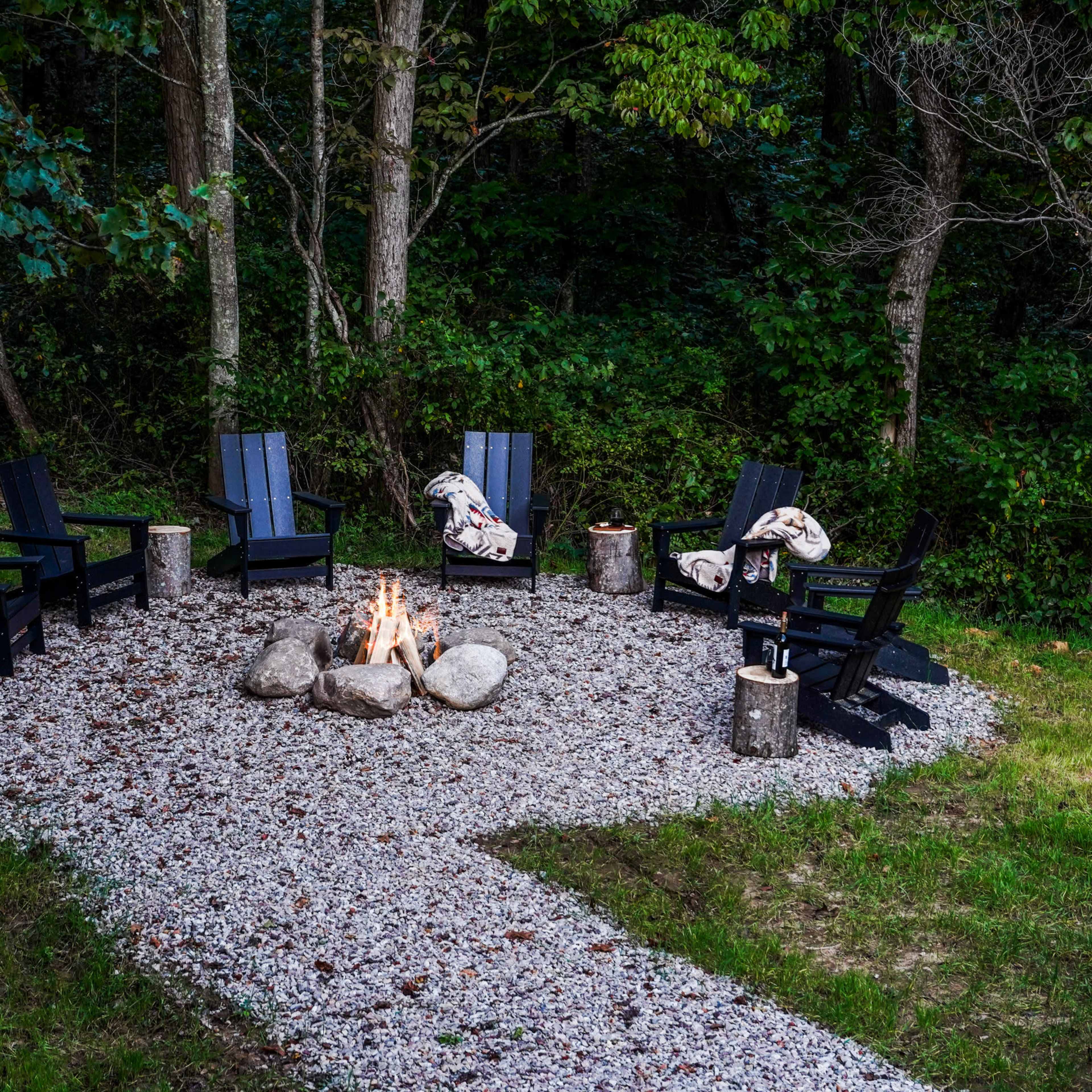 A circular stone fire pit area features six black chairs arranged around a central fire, surrounded by trees and gravel.