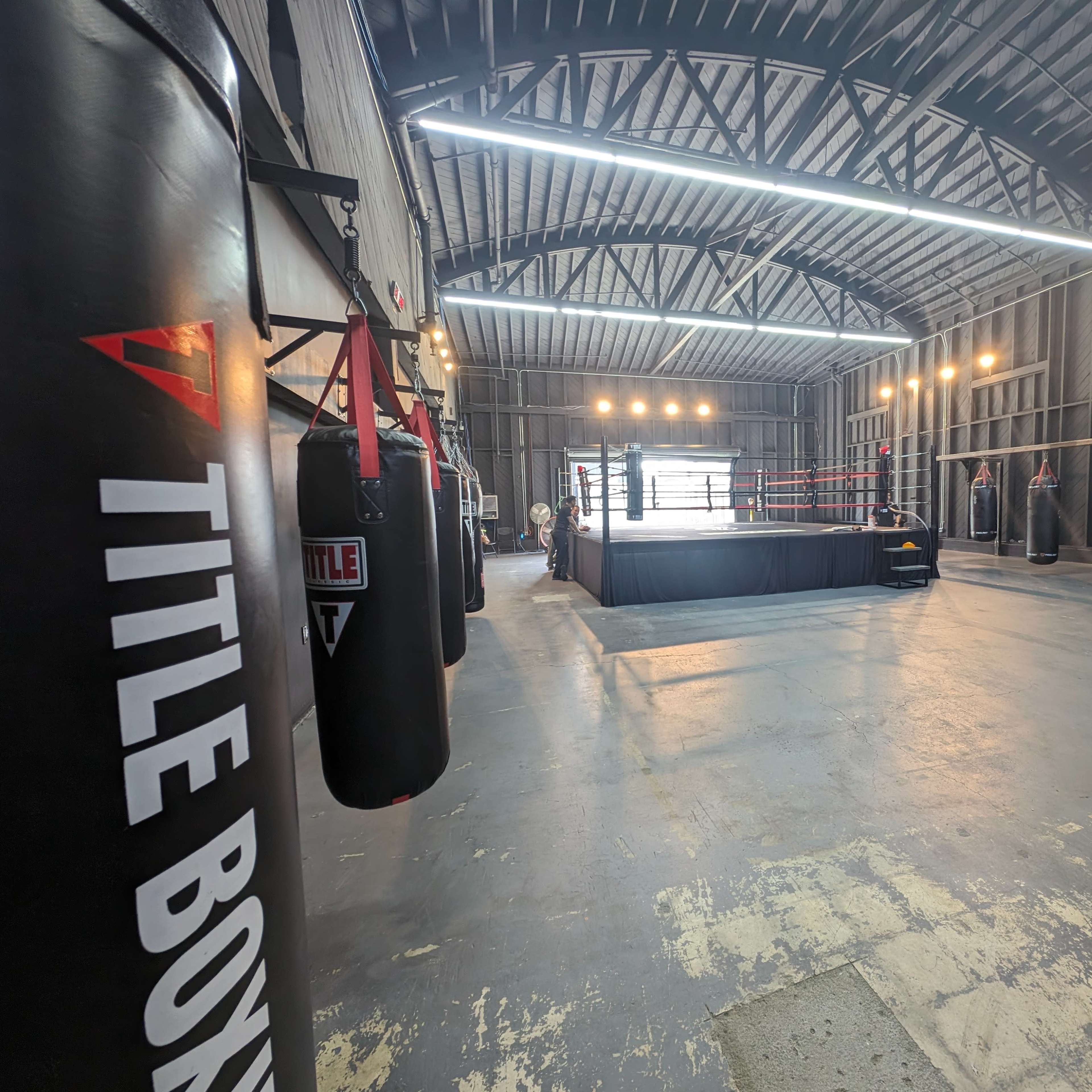 A spacious boxing gym featuring a central boxing ring surrounded by heavy punching bags and bright overhead lights.
