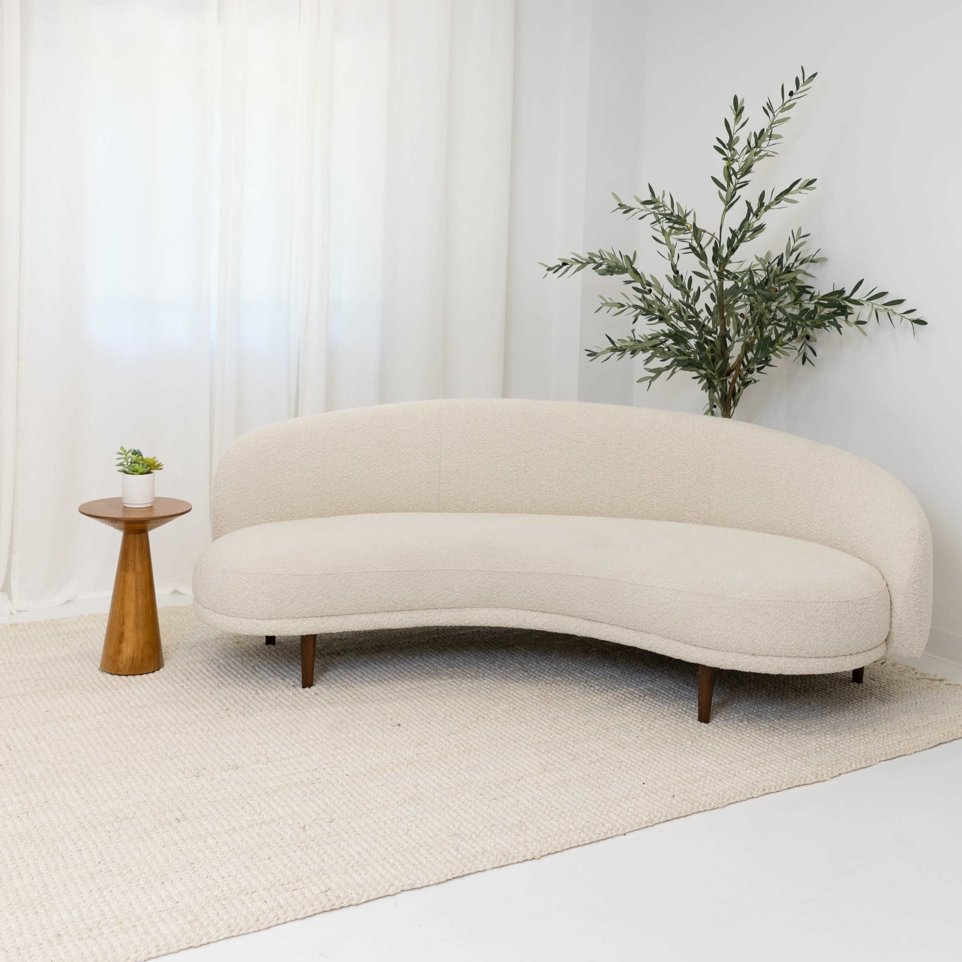 A curved beige sofa is positioned next to a small round table and a potted plant in a room with white walls and sheer curtains.
