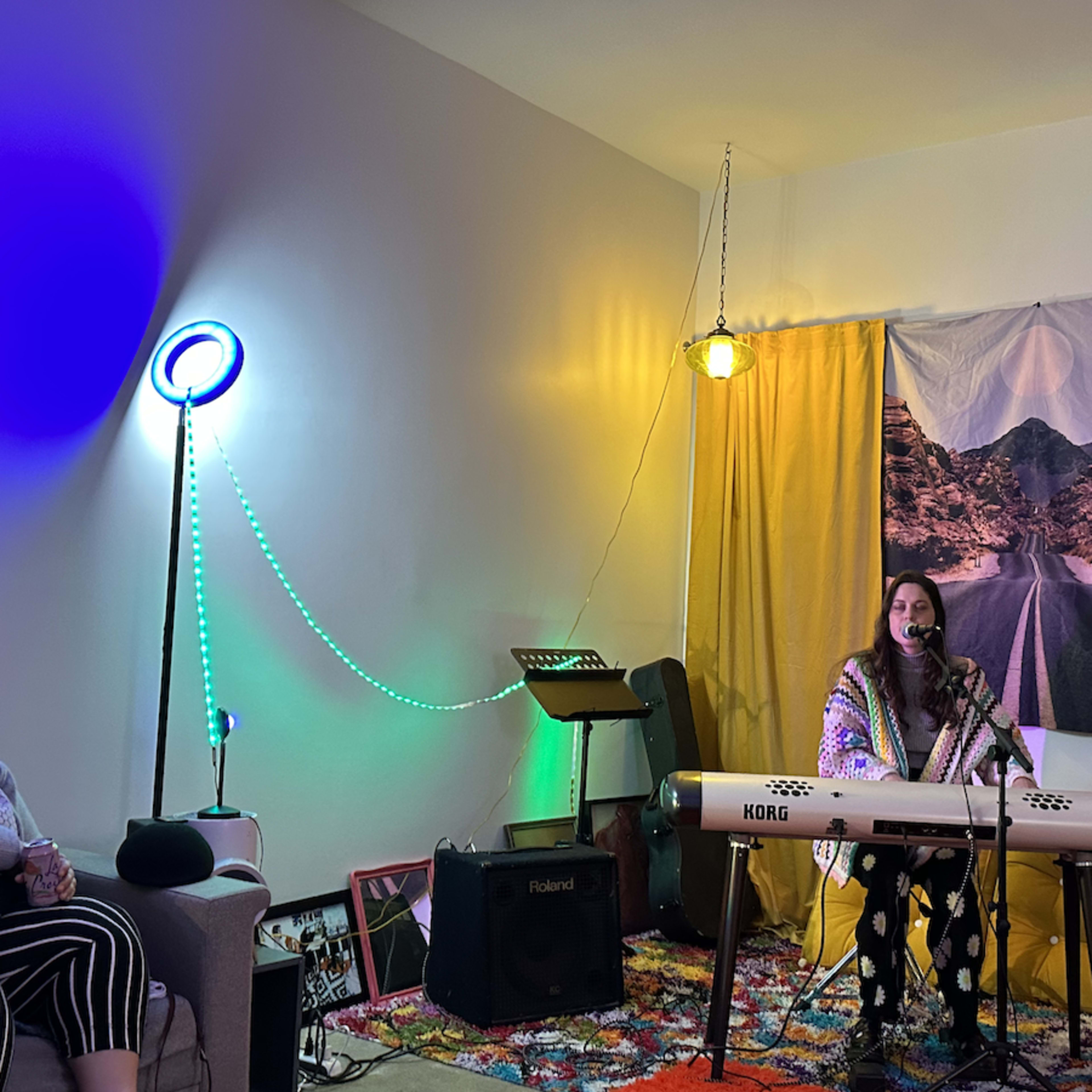 A woman sings and plays a keyboard in a cozy, colorful room while two audience members watch.