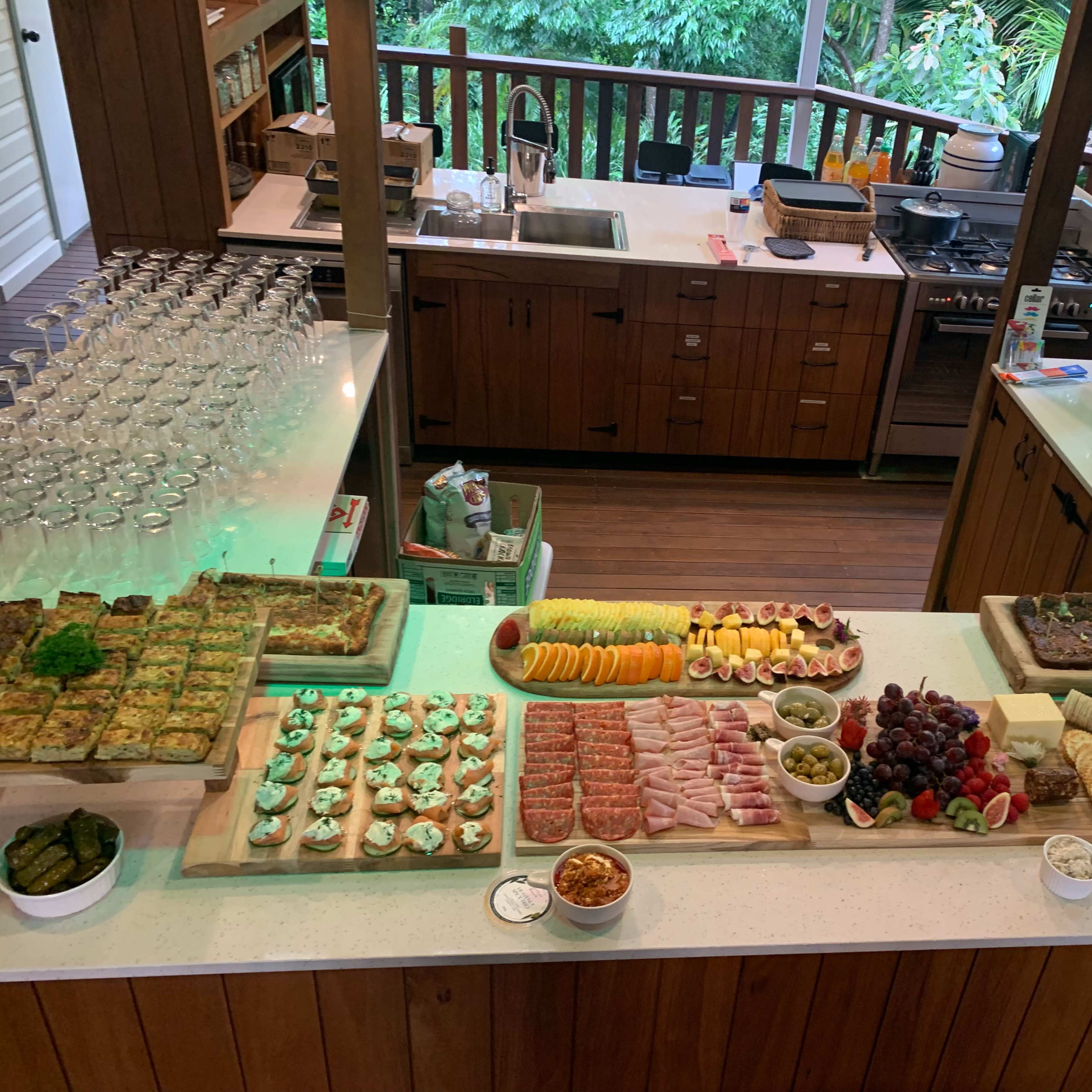 A large spread of food, including various fruits, appetizers, and desserts, is arranged on a countertop in a kitchen surrounded by greenery.