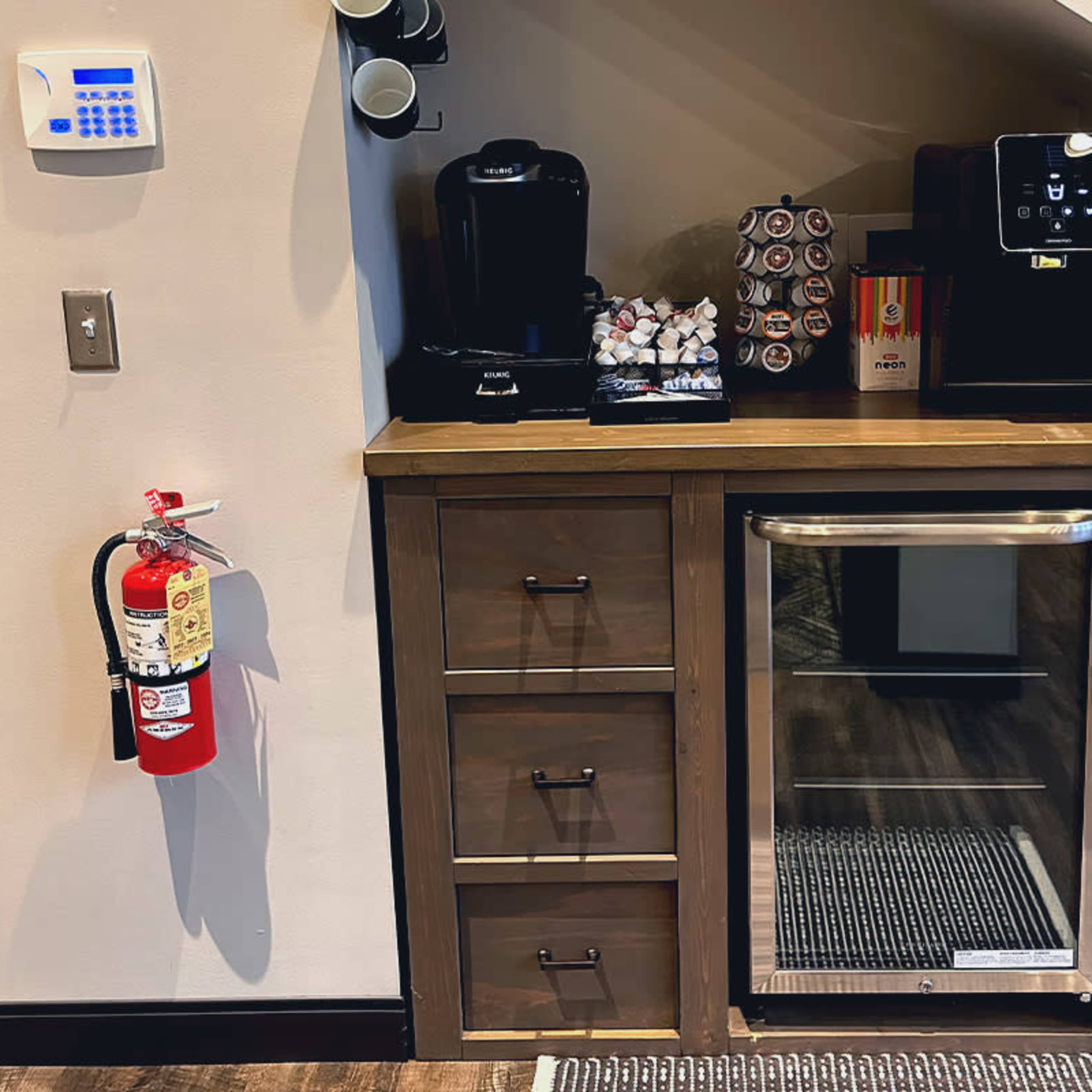 A kitchenette area featuring a coffee machine, a mini refrigerator, cabinets with three drawers, and containers of coffee pods on a wooden countertop.