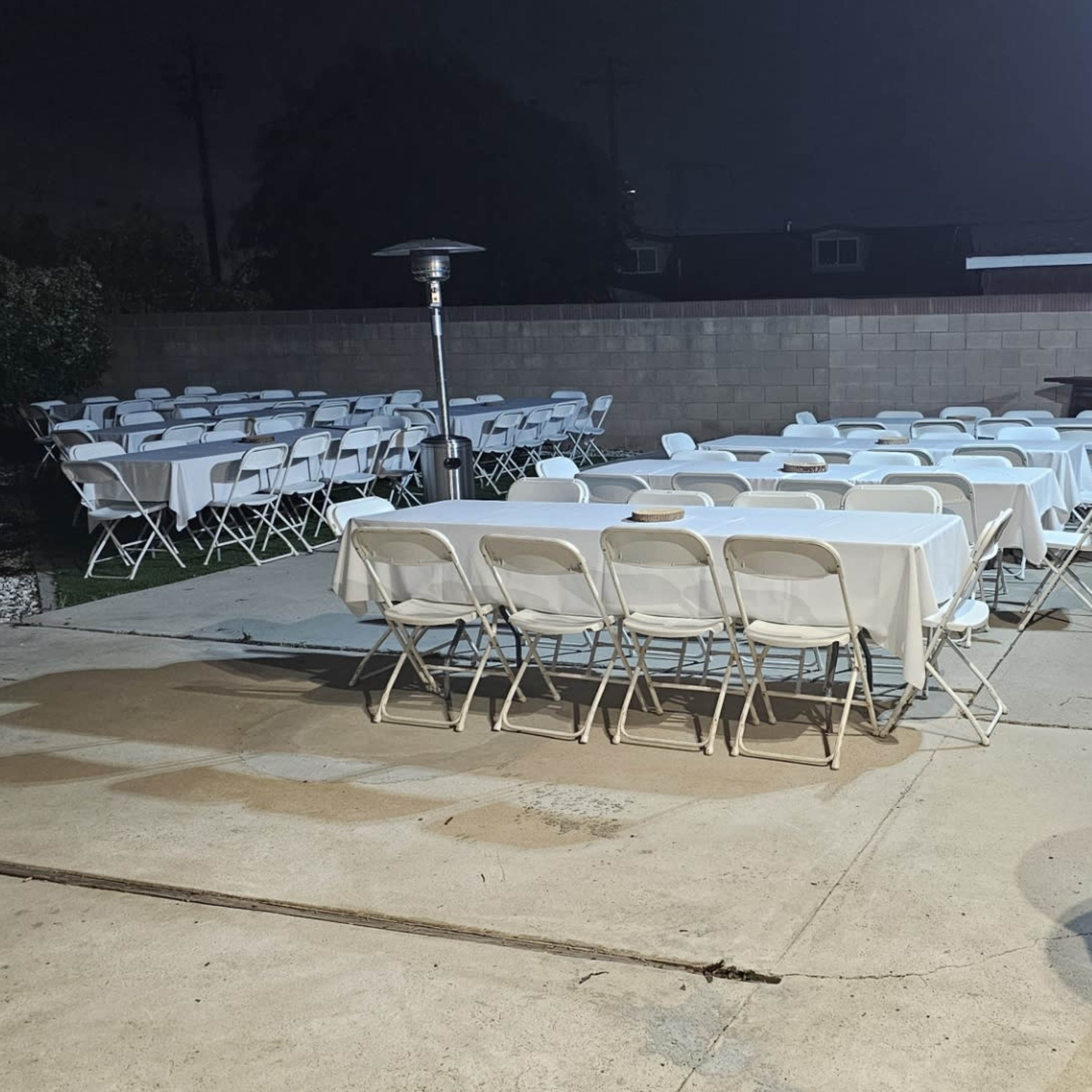 A row of white folding chairs is arranged around a patio area next to a wall, with a trash can in the corner and a heater in the background.