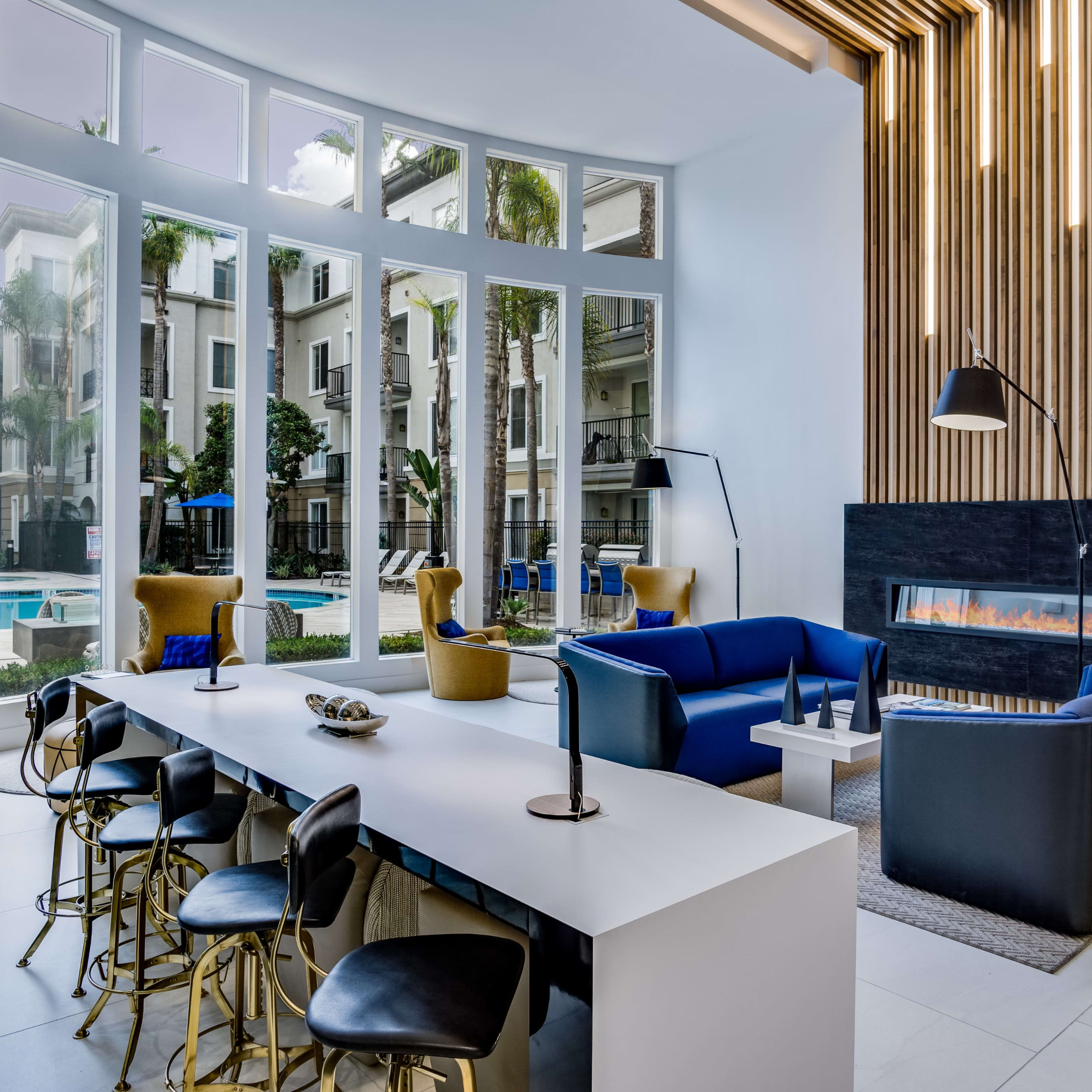 A modern indoor lounge area with a large table, stylish seating, and a fireplace, overlooking a pool surrounded by palm trees.