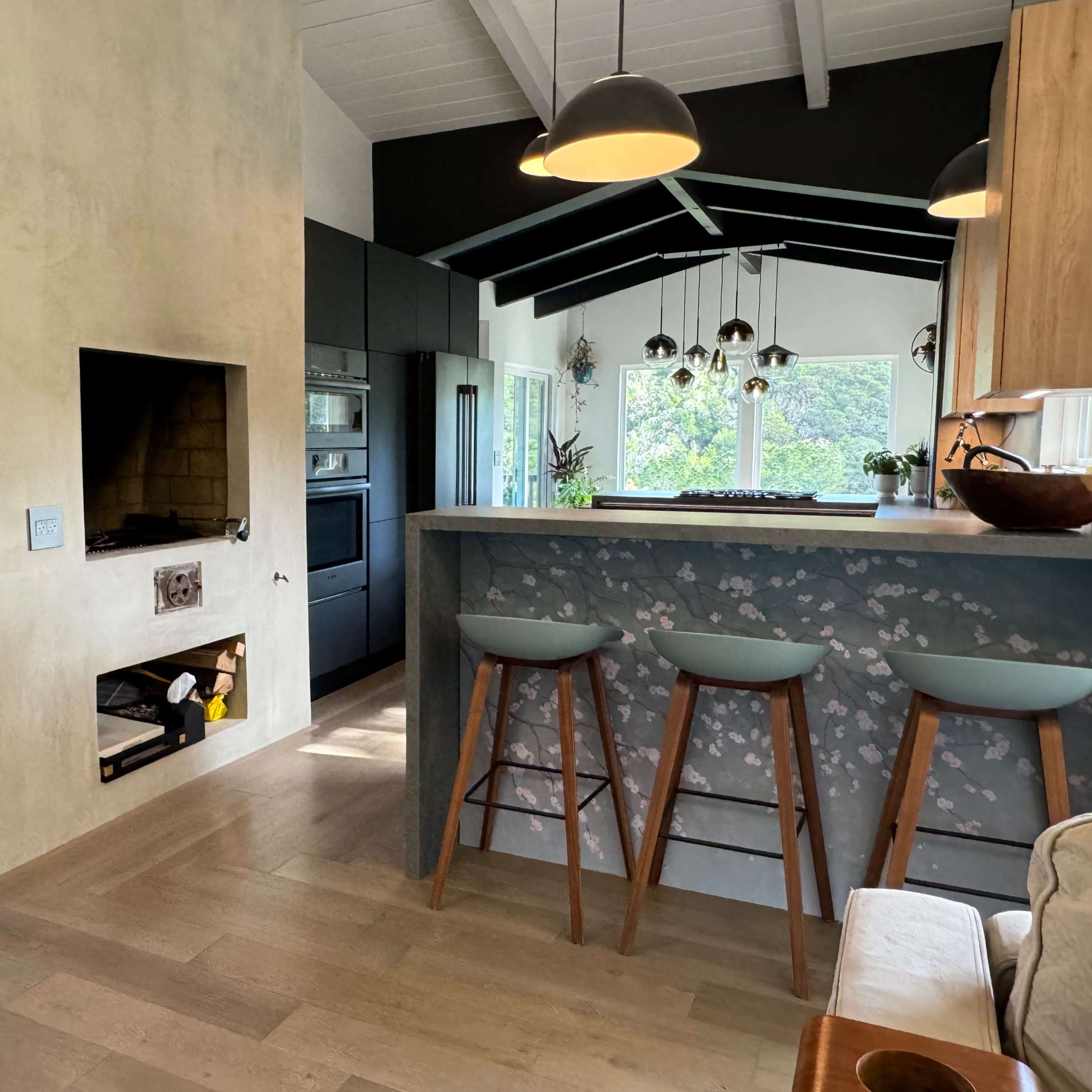 A modern kitchen with a bar area featuring three stools, a built-in fireplace, and large windows allowing natural light to enter.