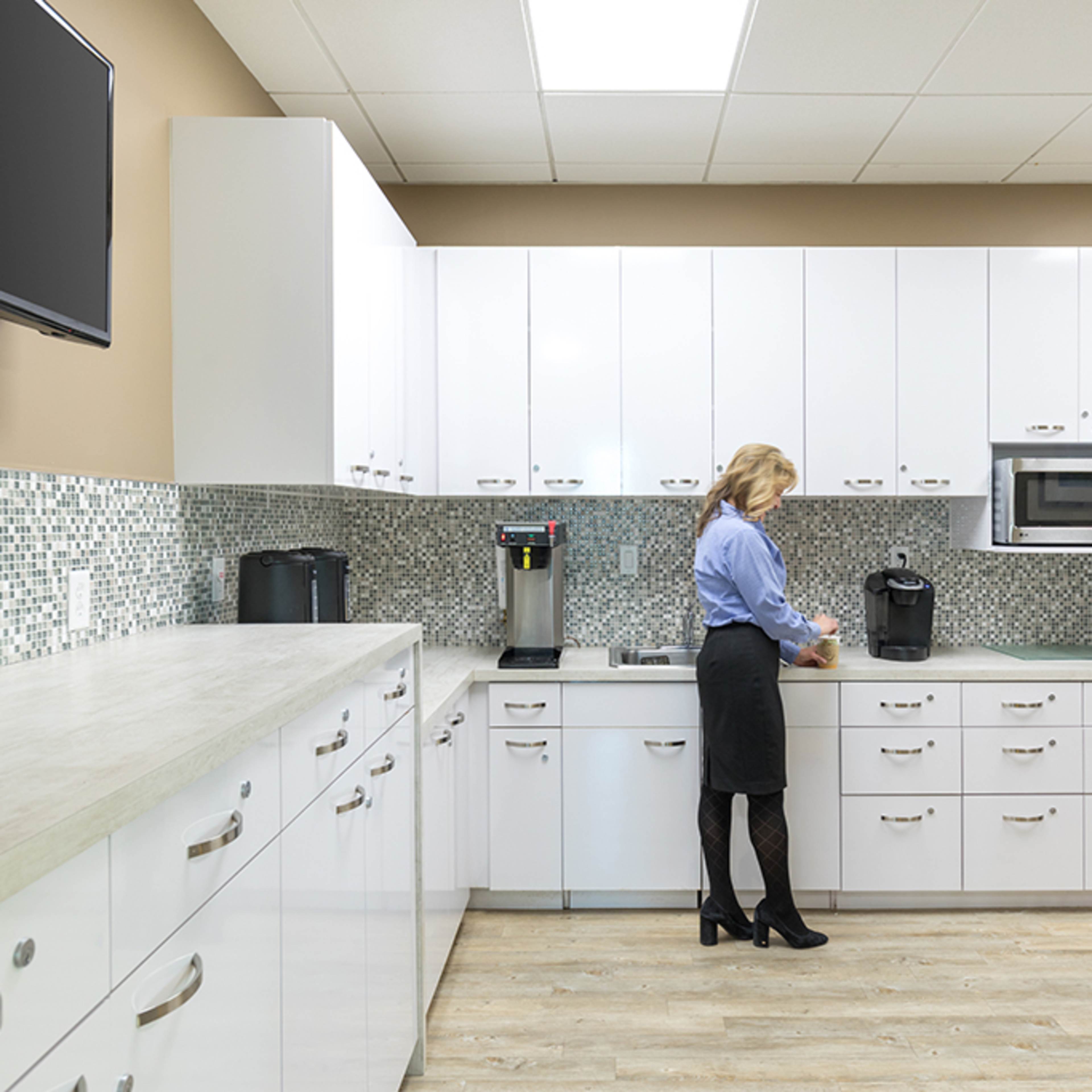 A woman in a blue shirt prepares coffee in a modern kitchen with white cabinets and stainless steel appliances.