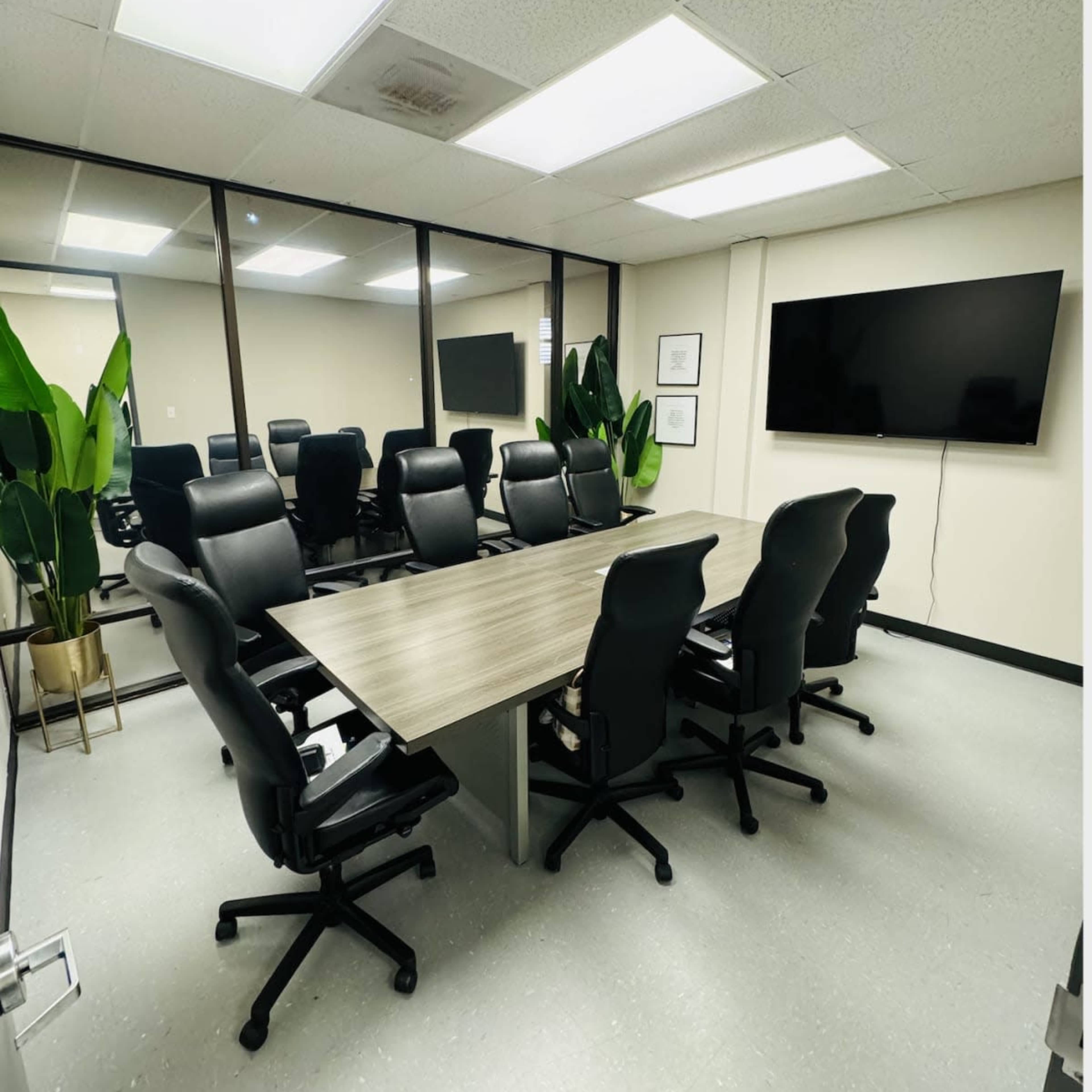 A modern conference room with a long table surrounded by black office chairs, and large windows facing a wall-mounted television.