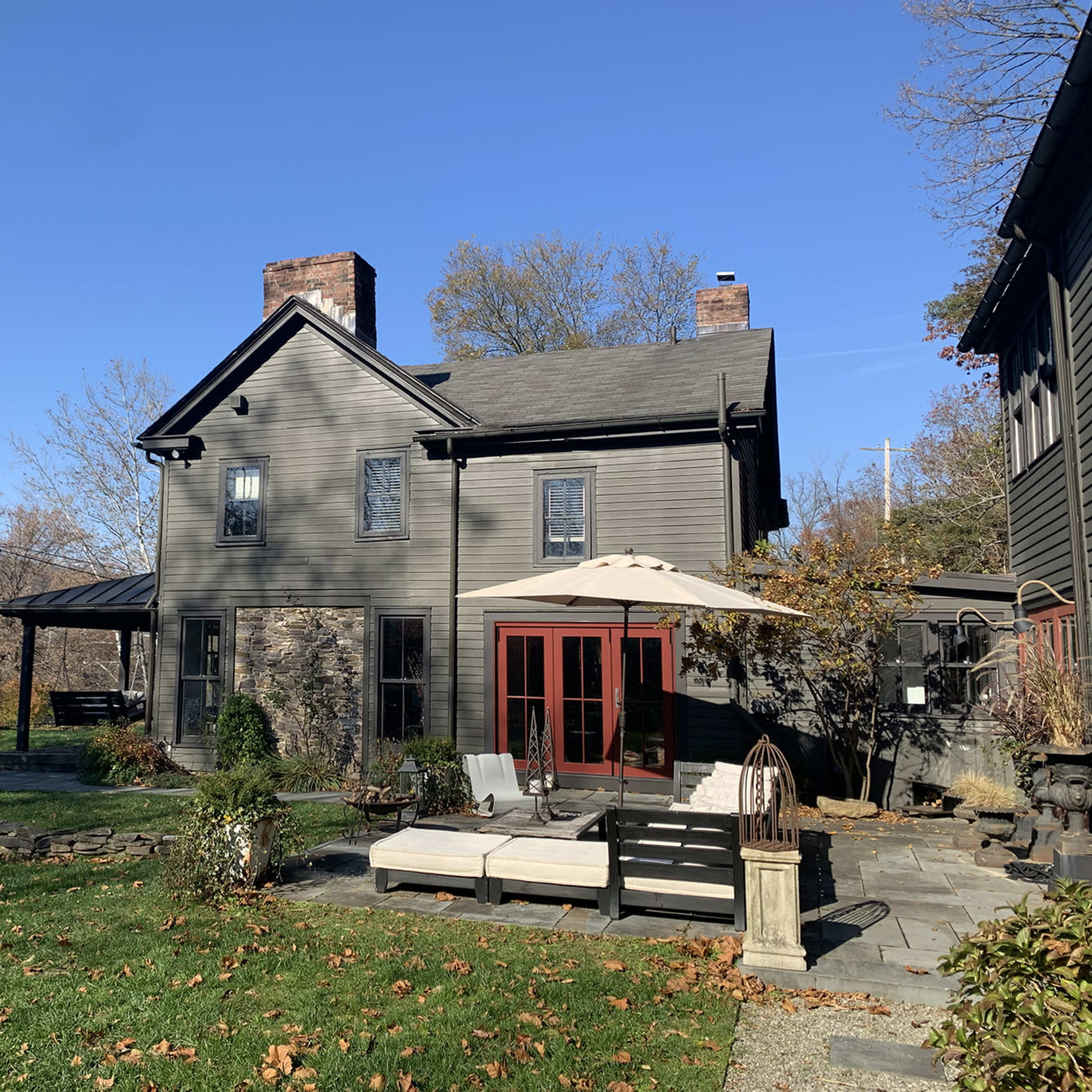 A two-story gray house with a patio and outdoor seating overlooks a river, surrounded by trees and a lawn.