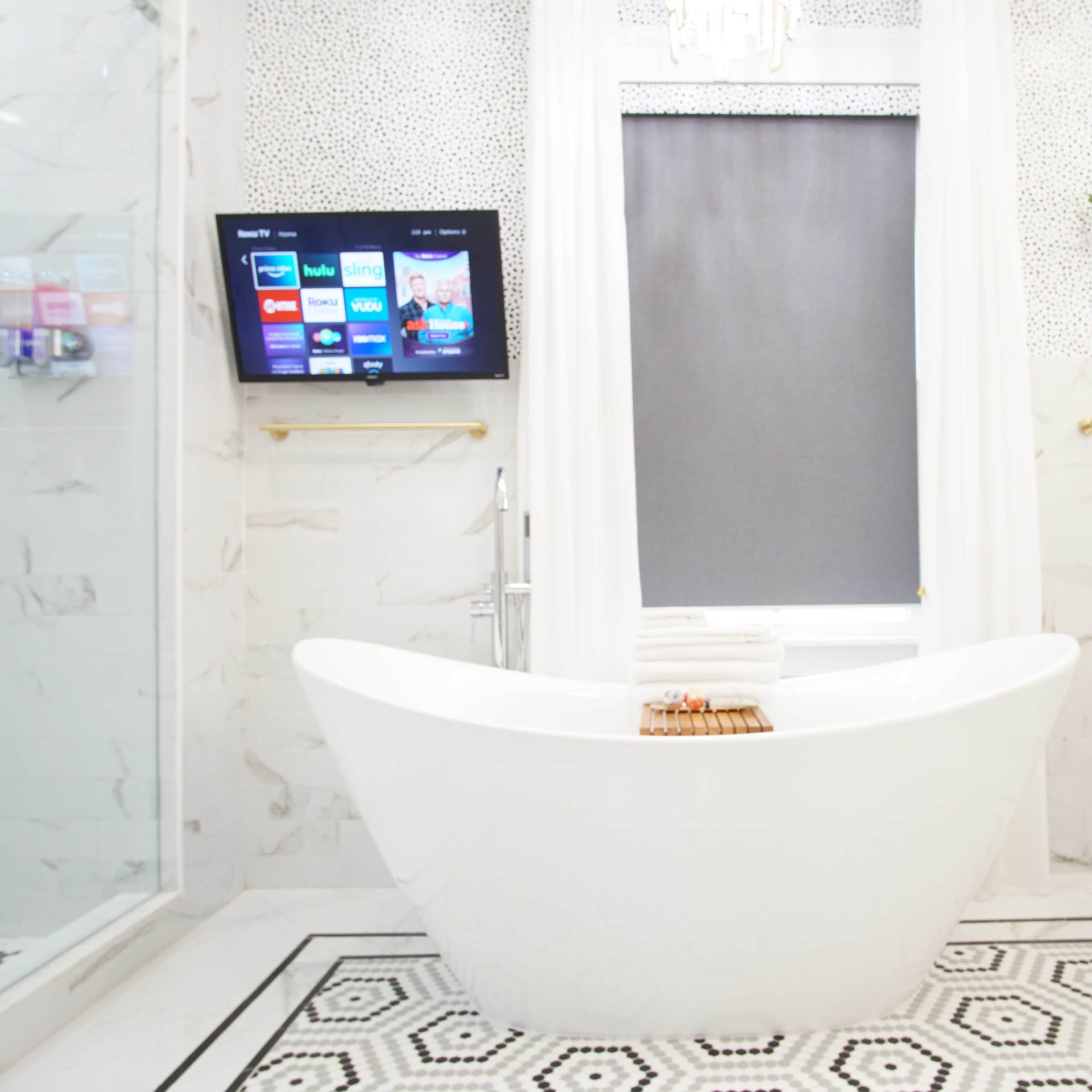 The bathroom features a freestanding white bathtub, a wall-mounted television, and a hexagonal patterned rug.