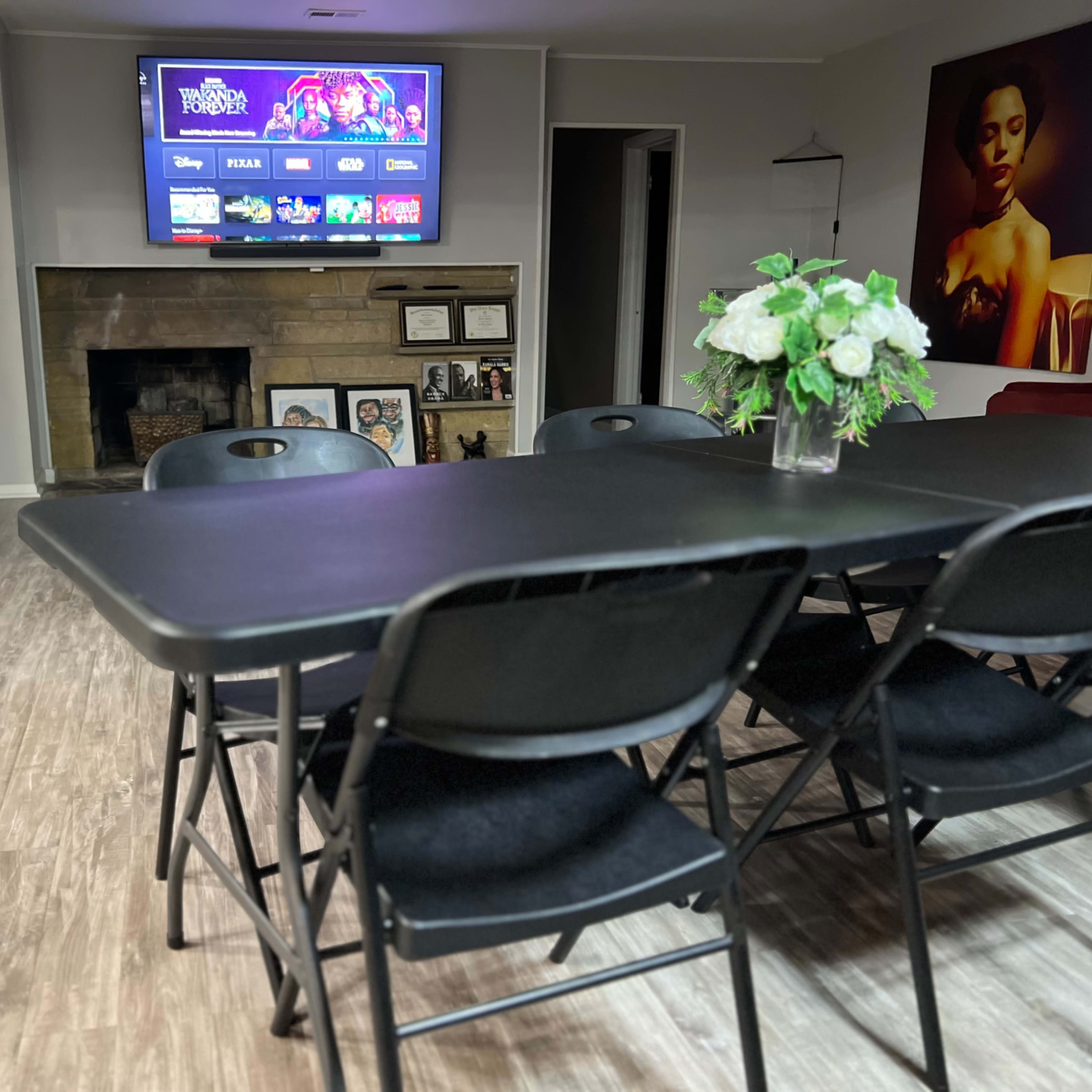 A room with a black folding table and chairs arranged in front of a television mounted on the wall, showcasing a selection of apps, with a fireplace and artwork visible in the background.