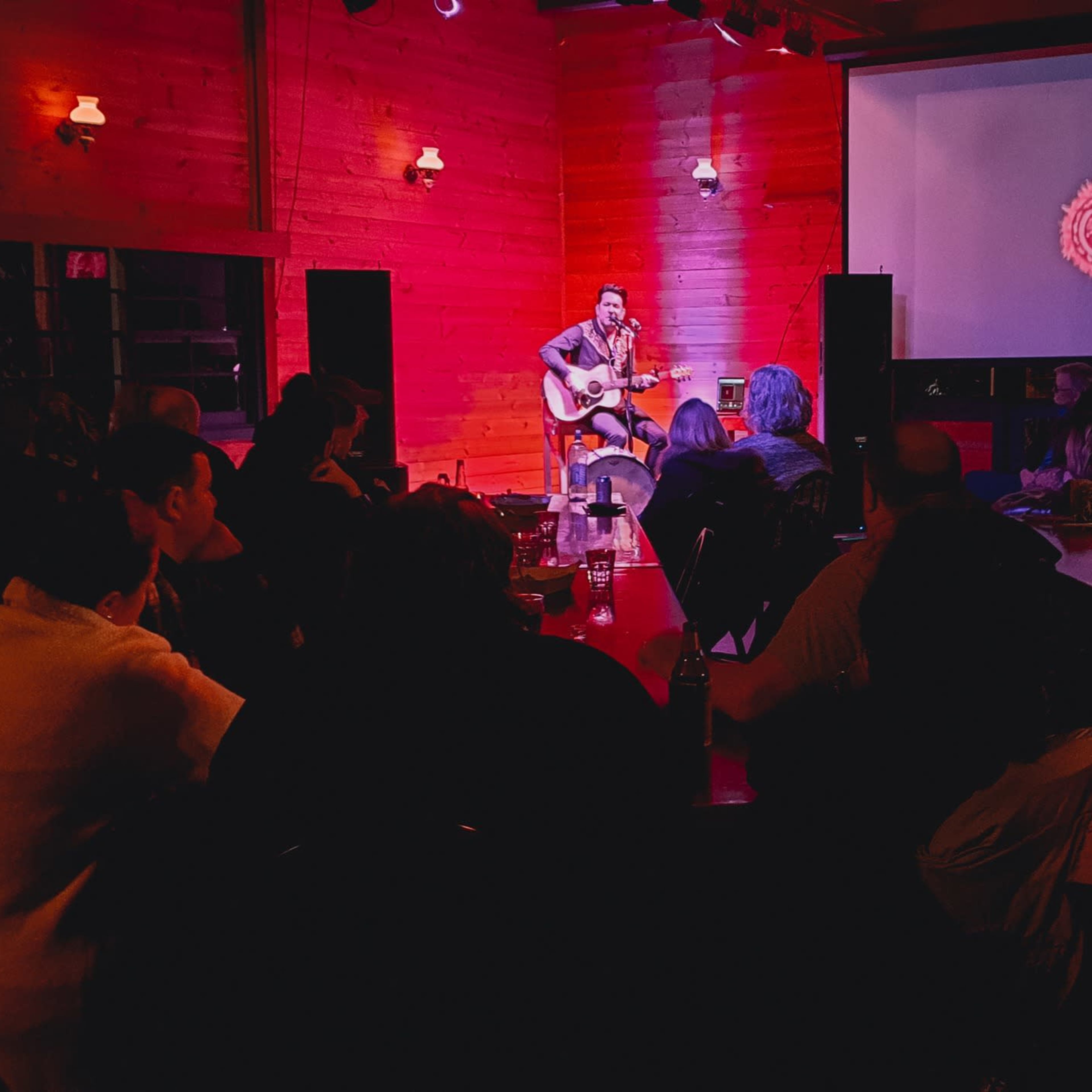 A musician plays guitar on stage in a dimly lit venue with an audience seated at tables.