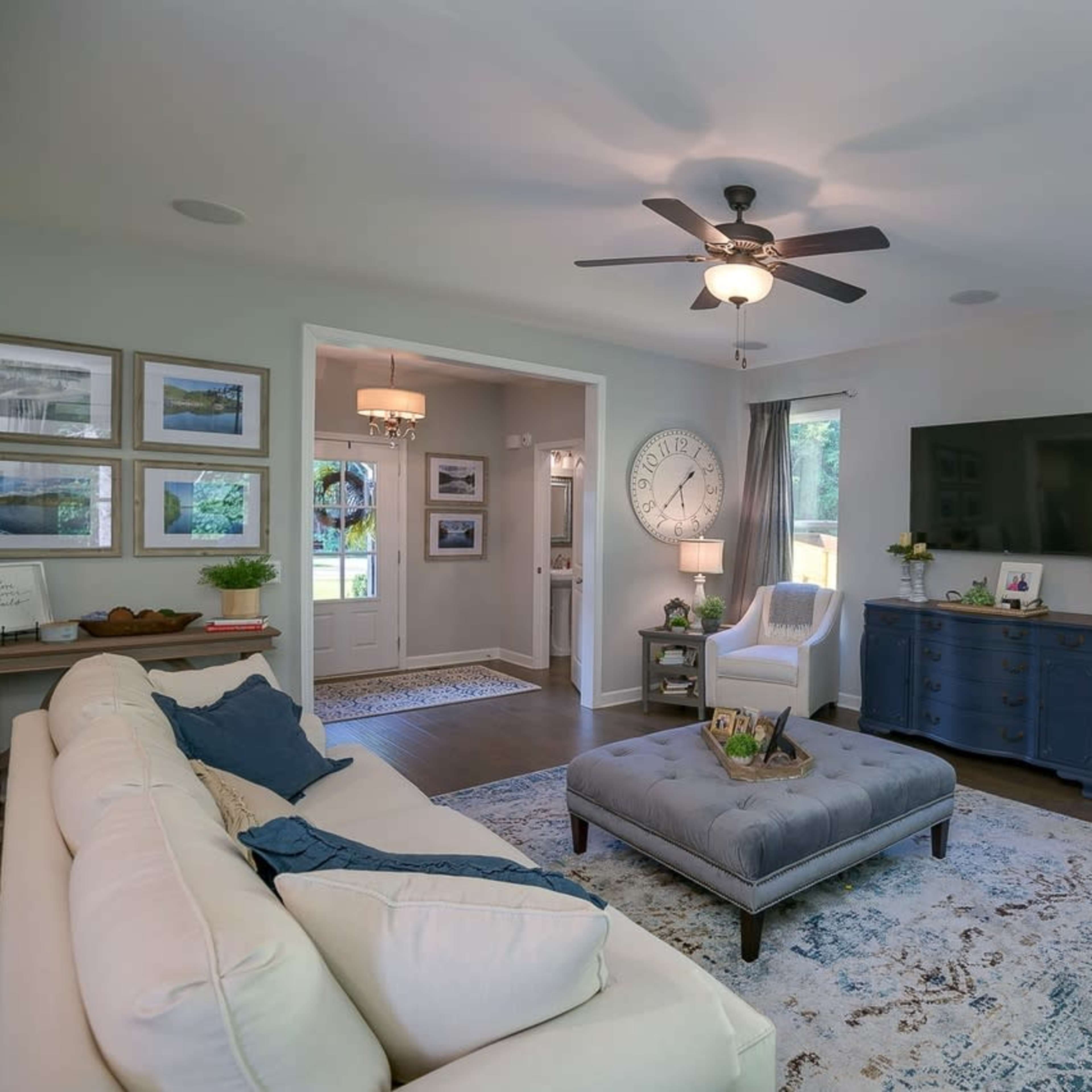 A modern living room with a beige sofa, blue ottoman, a dark wooden console table, and framed pictures on the walls, all illuminated by natural light from windows.