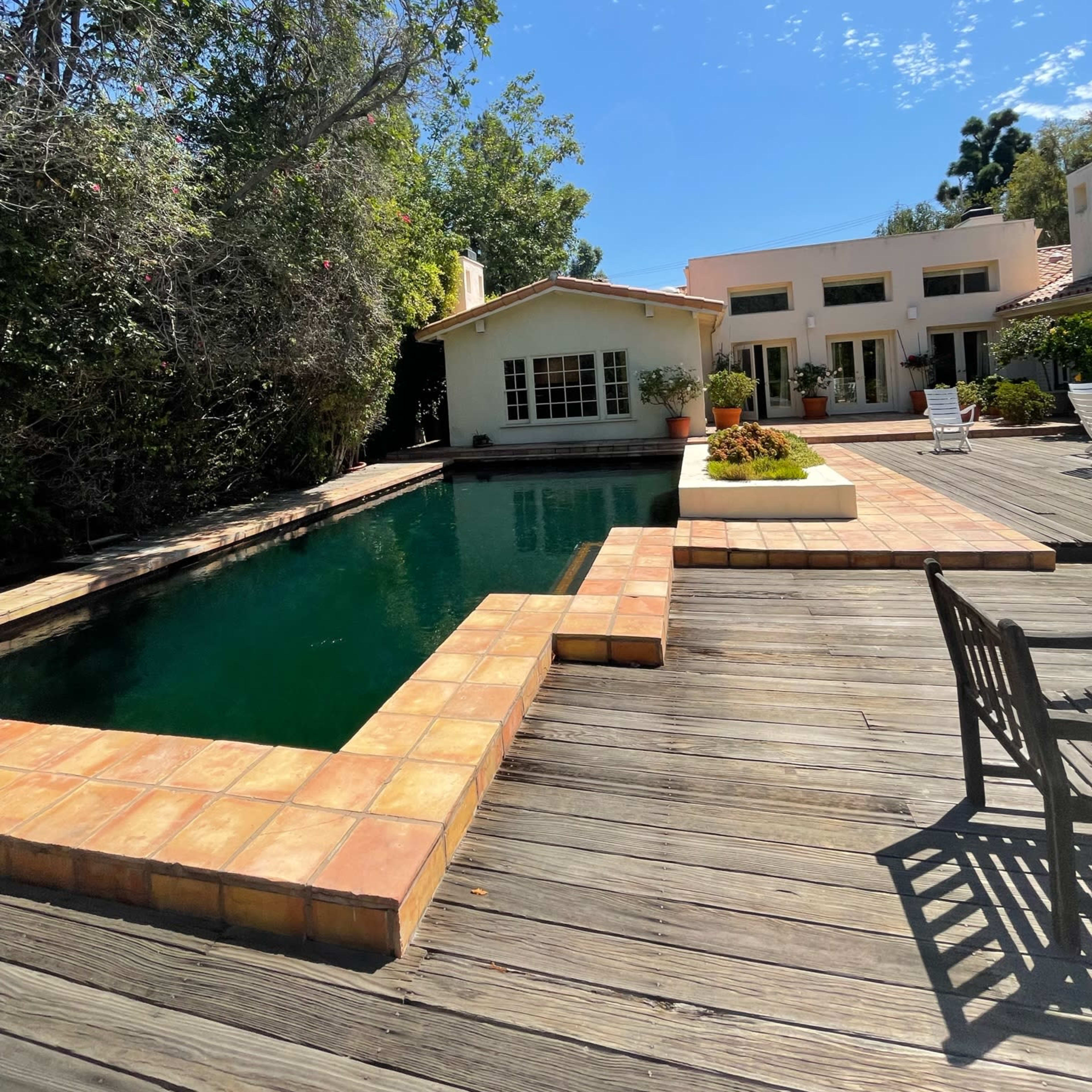 A swimming pool with a tiled border is surrounded by wooden decking and a garden, with a house visible in the background under a clear blue sky.