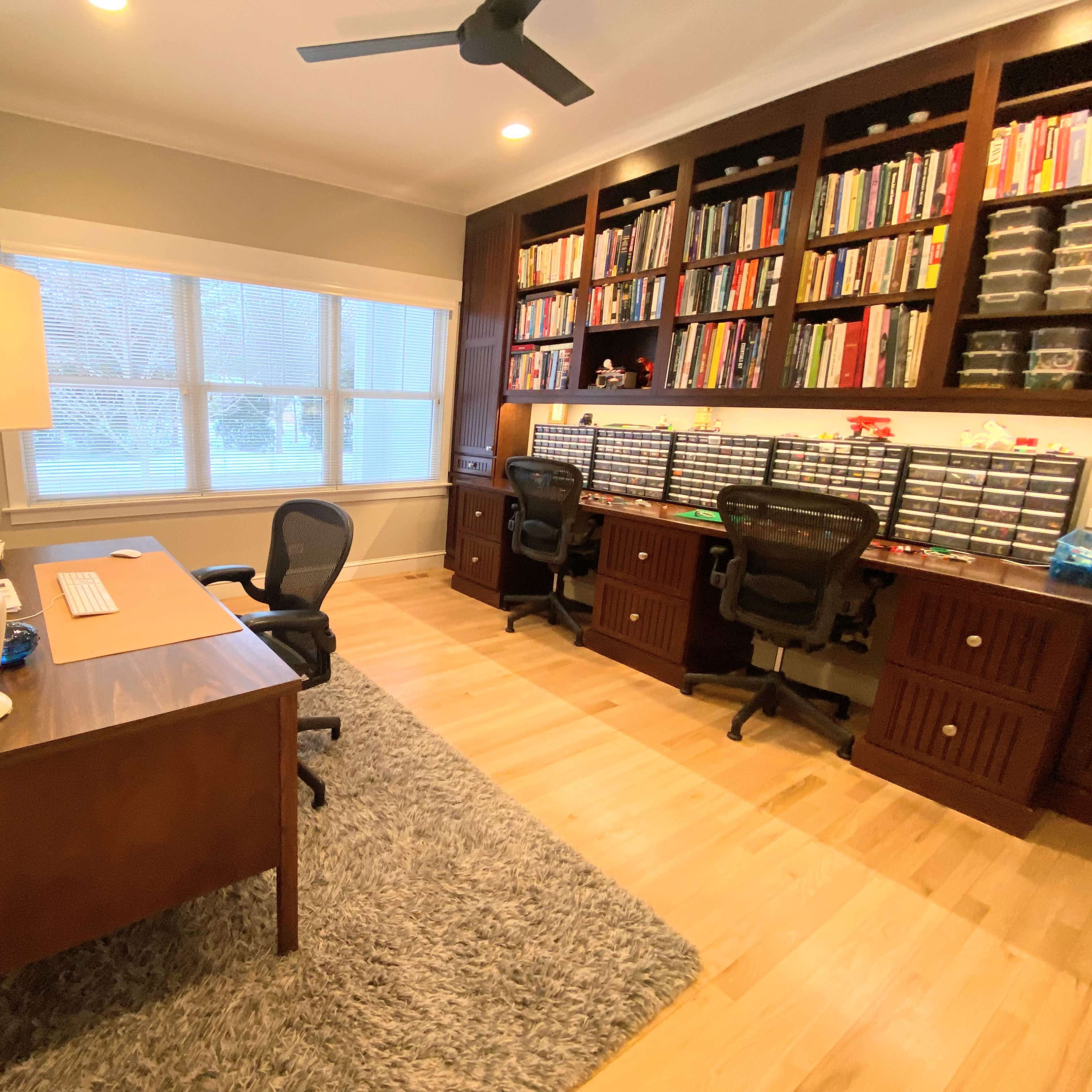 A home office features a wooden desk with a lamp, two office chairs, and a wall of bookshelves filled with books and storage containers.