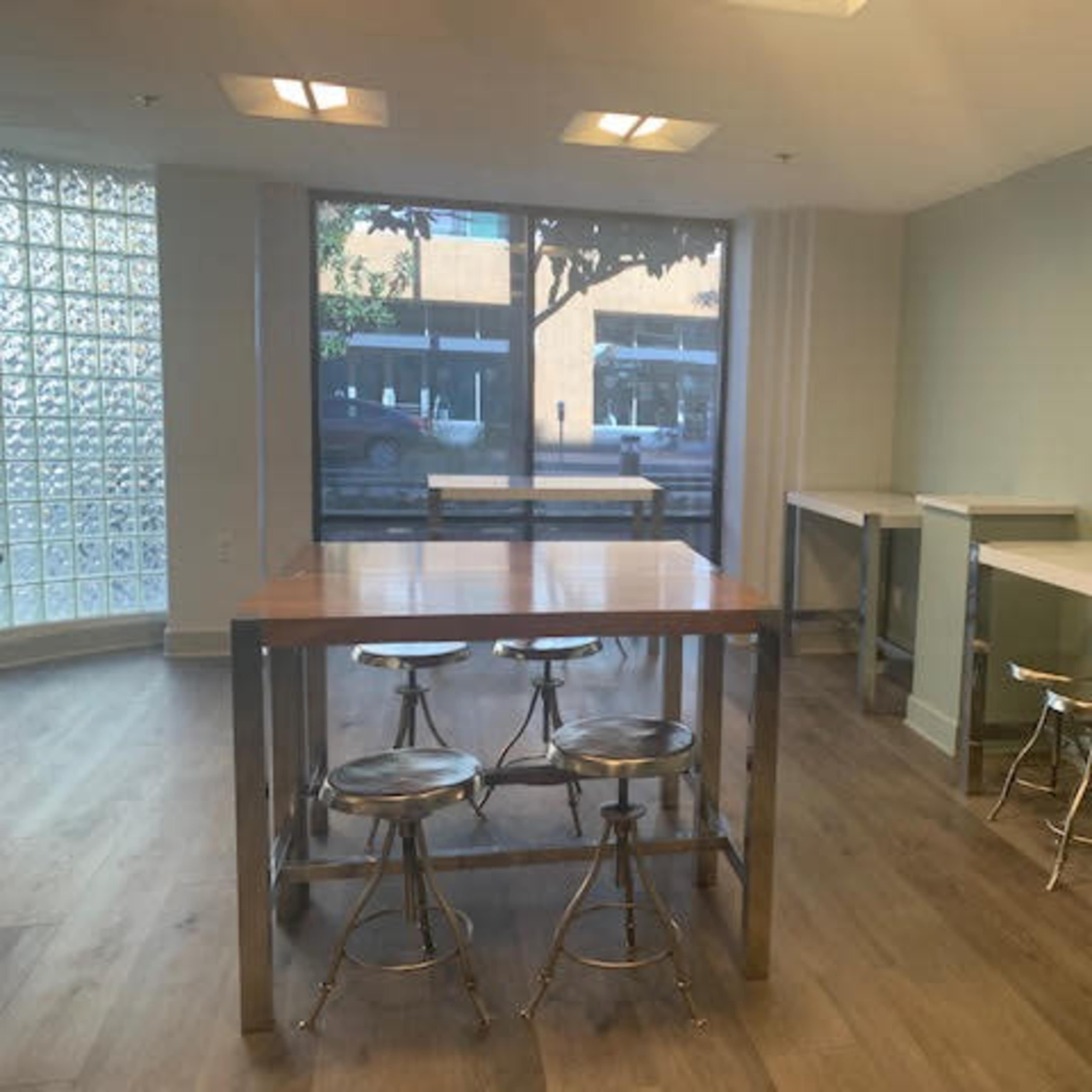 A room with several tall, rectangular tables and metal stools, featuring large windows that let in natural light.