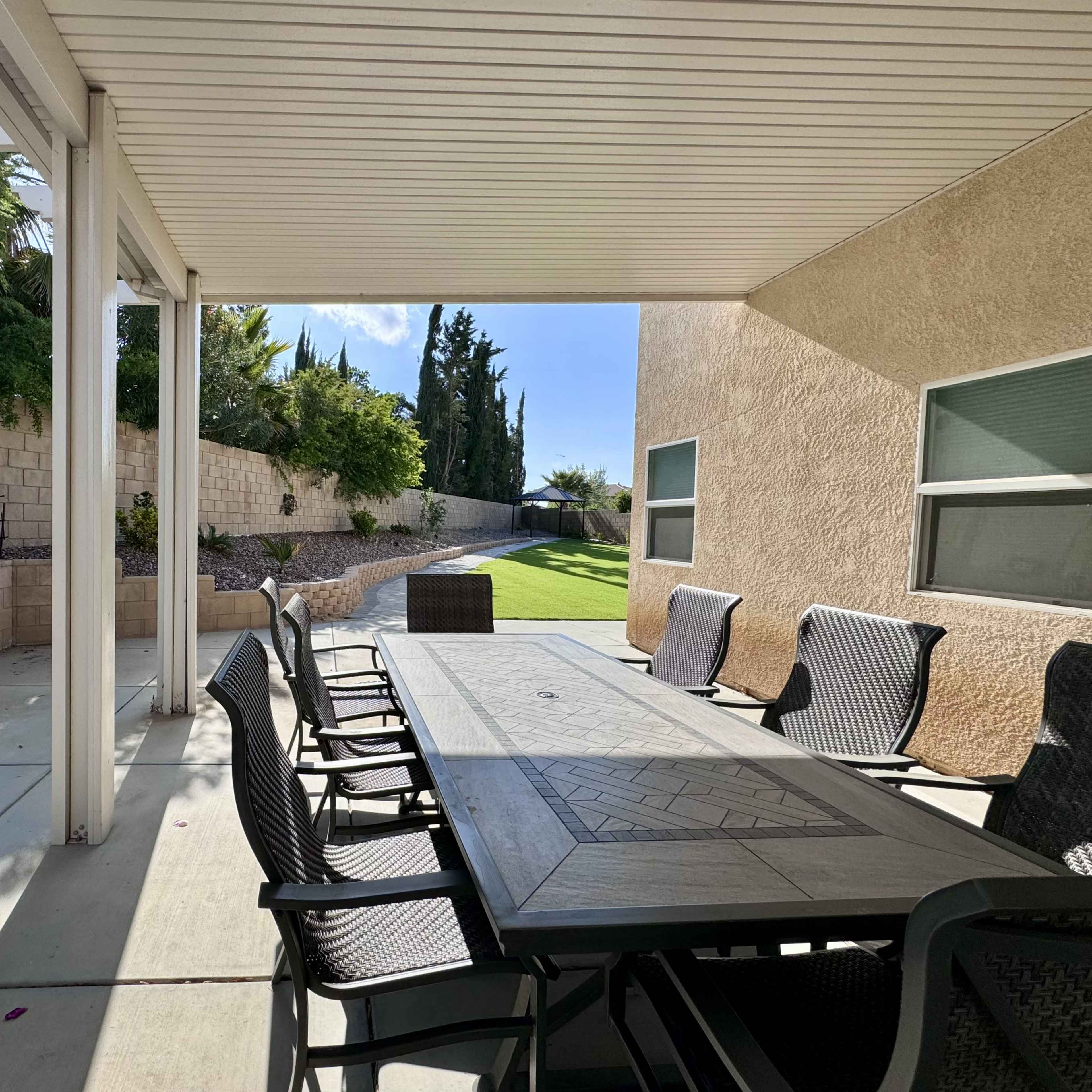 A shaded outdoor dining area features a large table surrounded by black chairs, overlooking a grassy yard with shrubs and trees in the background.