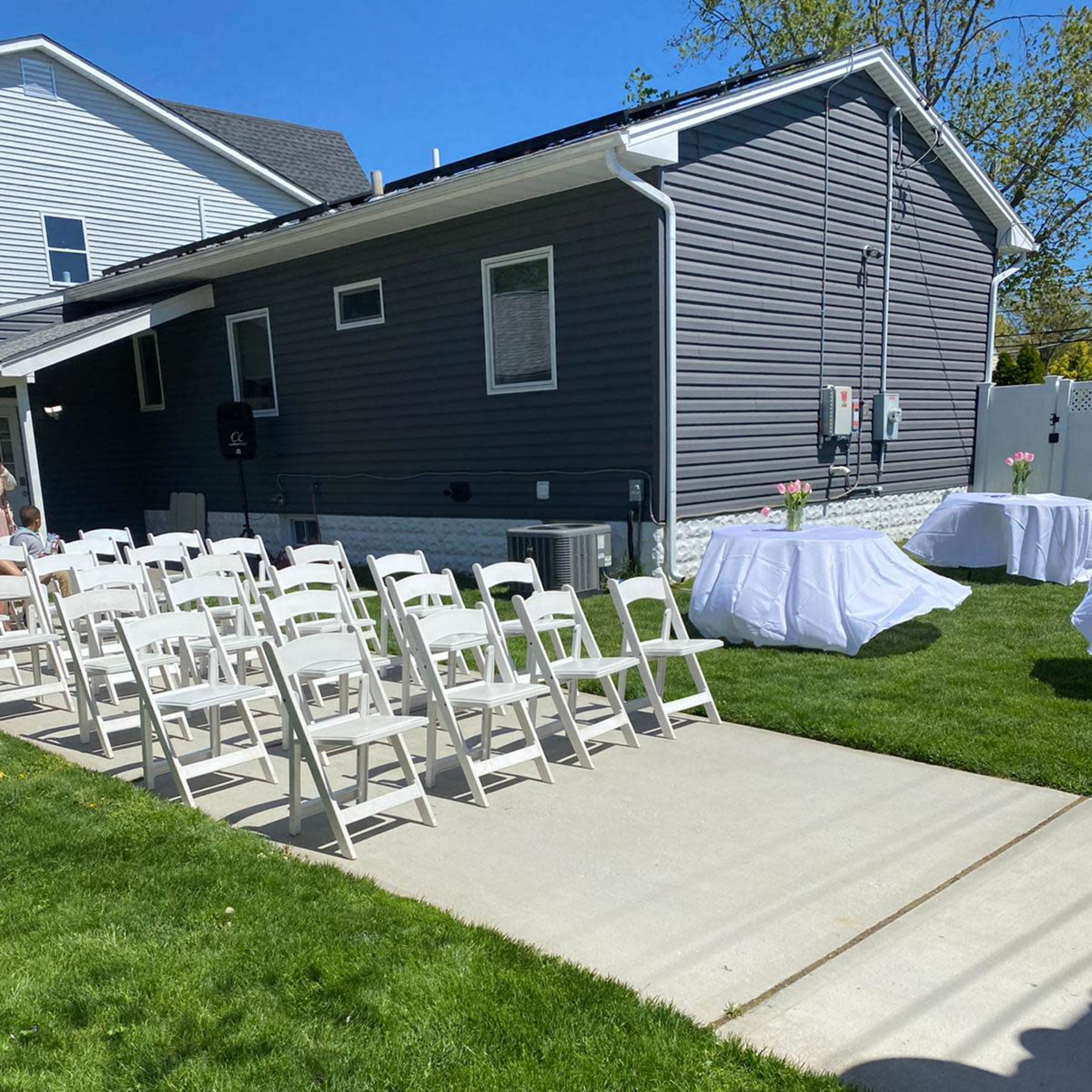 A row of white folding chairs is neatly arranged on a concrete path beside a grassy area next to a gray house, with a table covered in white cloth in the background.