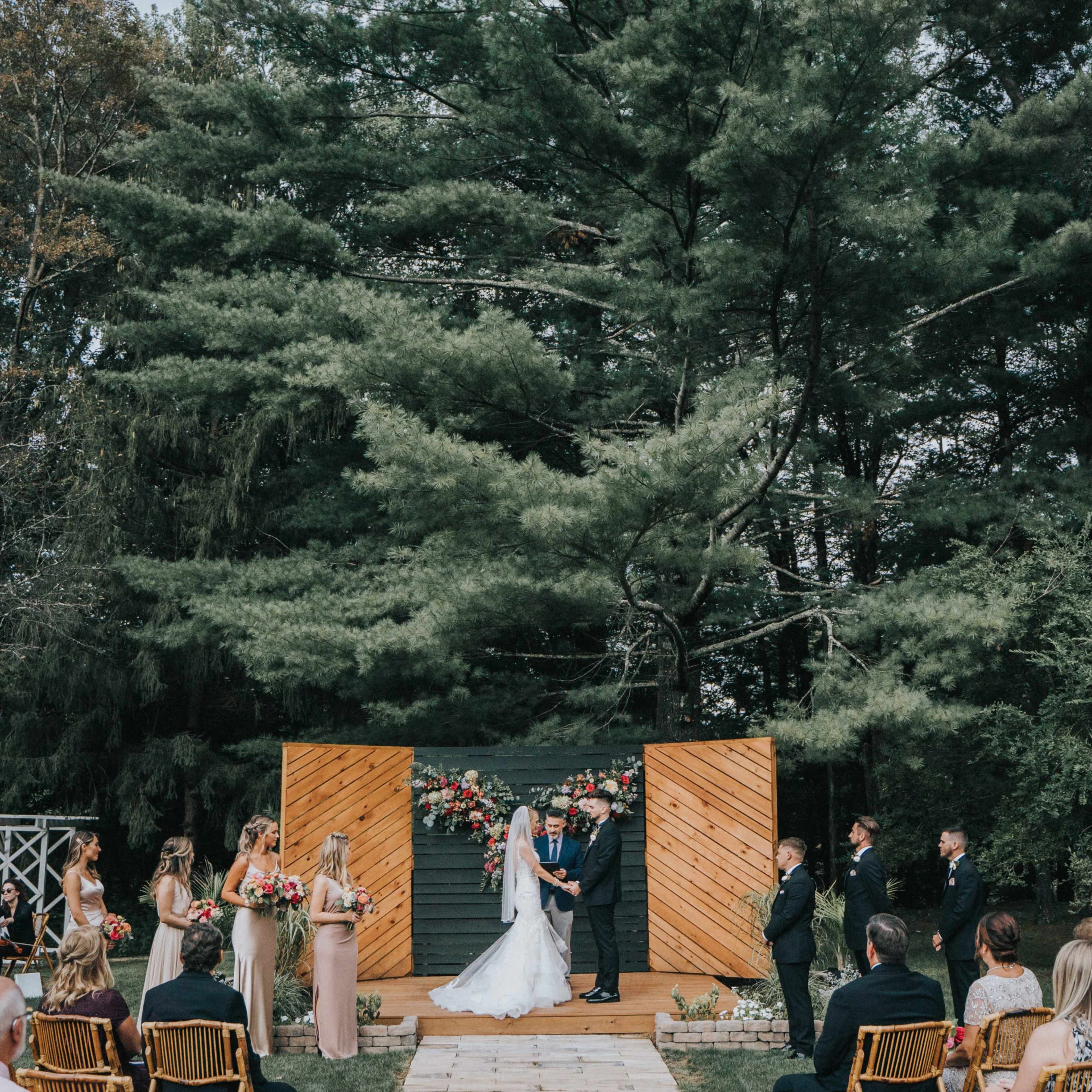 A wedding ceremony takes place outdoors on a wooden platform, surrounded by guests seated in rows and flanked by trees.