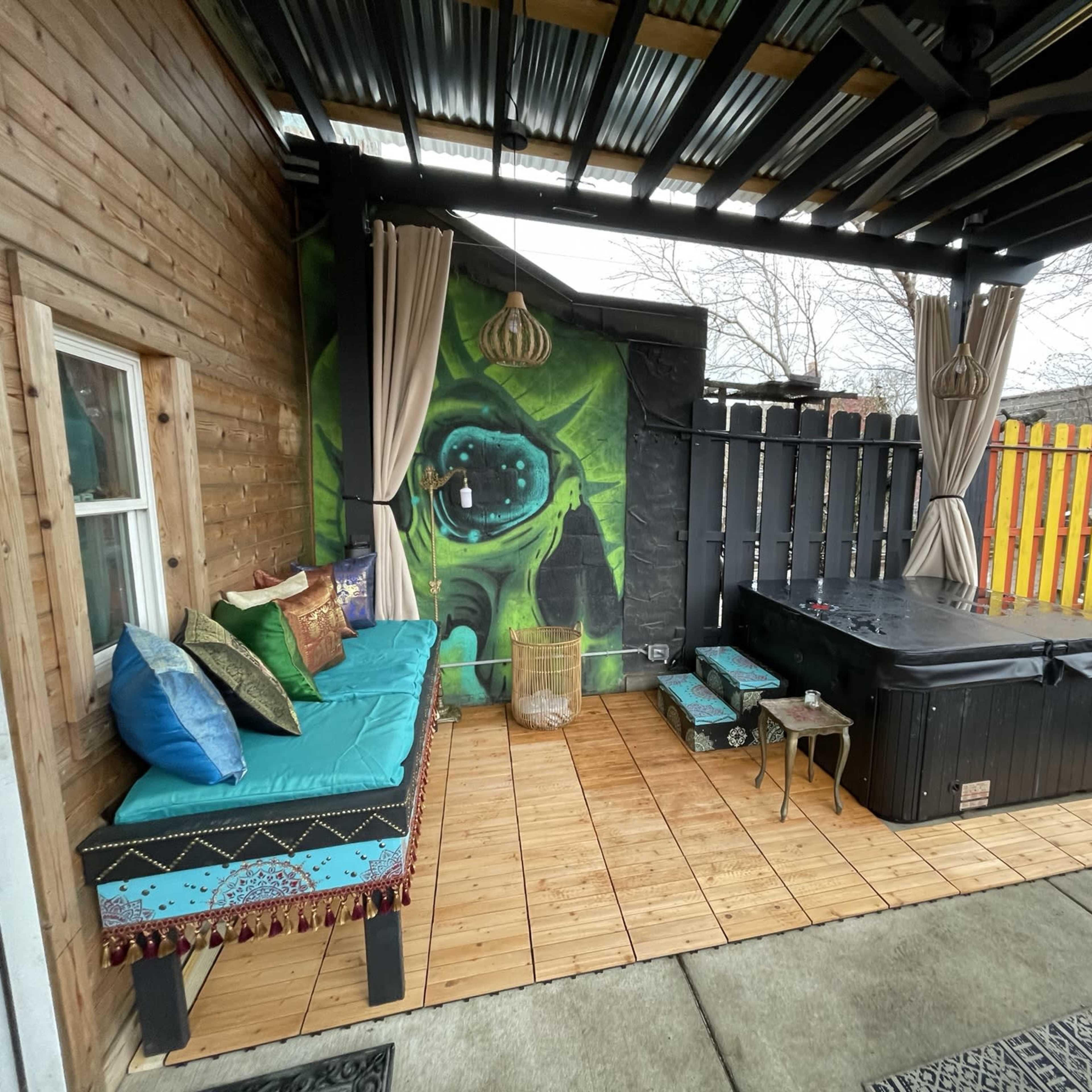 A covered outdoor patio area with a spa, wooden flooring, a wall mural, and cushioned seating.