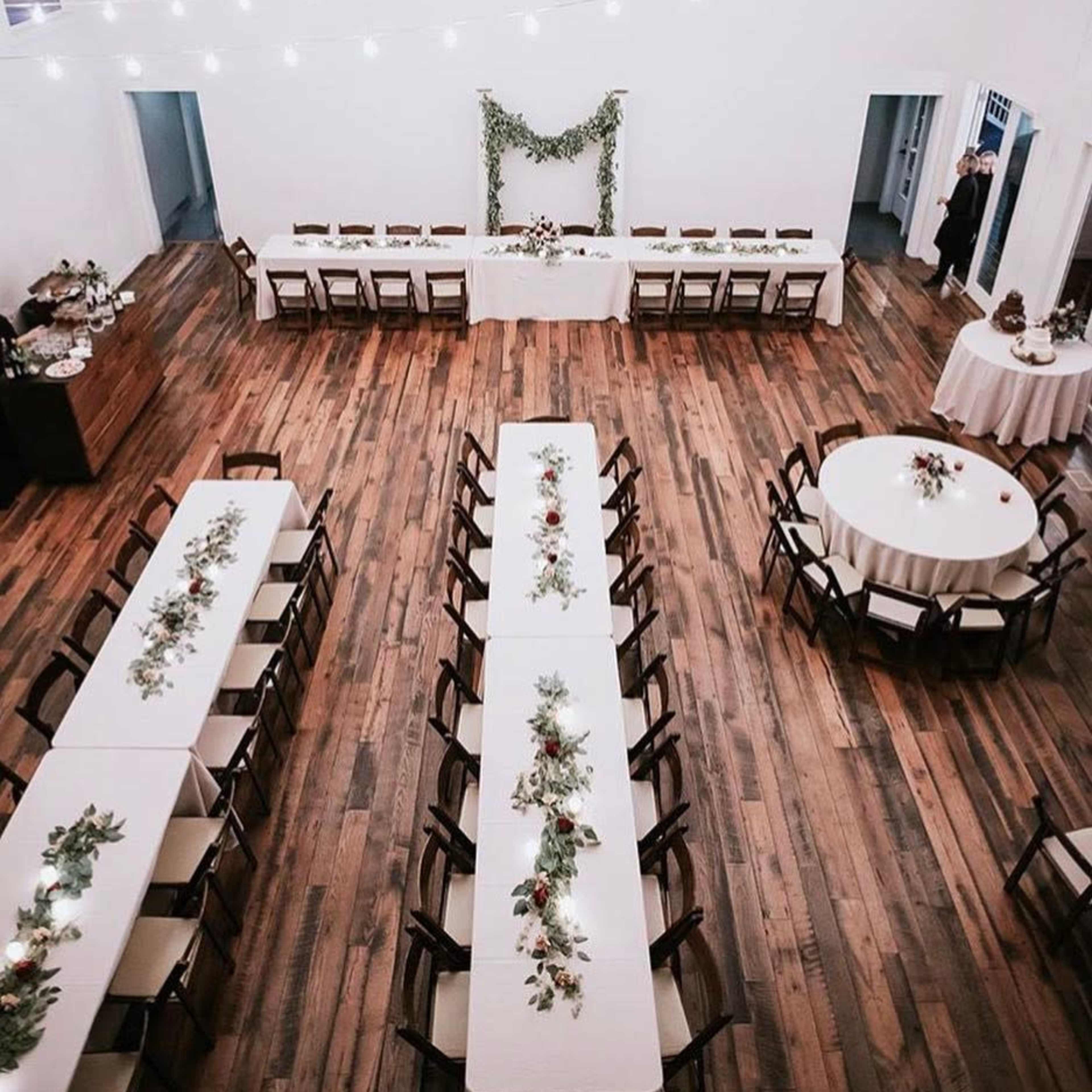 A spacious indoor venue features multiple long dining tables arranged with decorative centerpieces and string lights overhead.