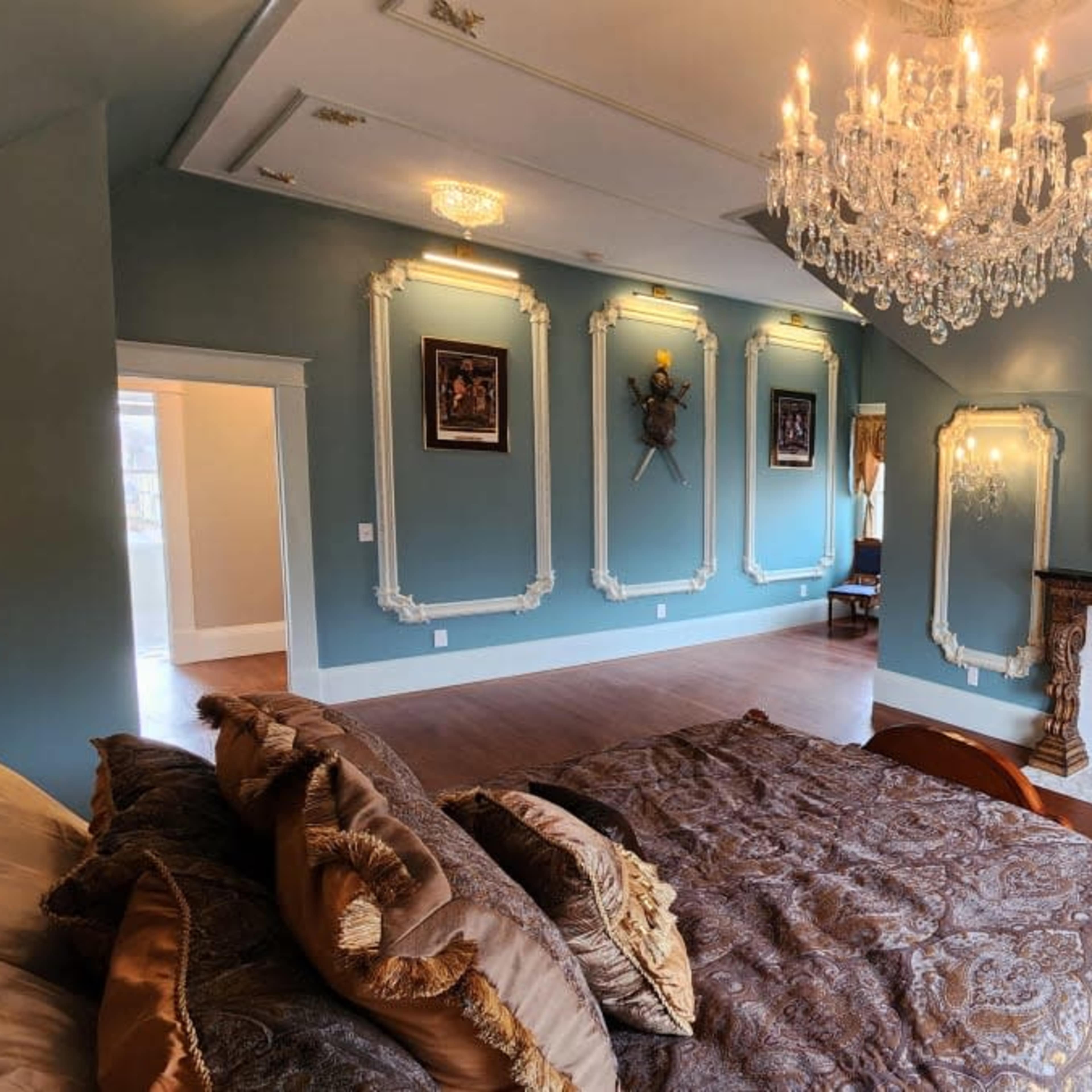 A spacious bedroom features a crystal chandelier, ornate wall decor, and a fireplace surrounded by elegant molding.