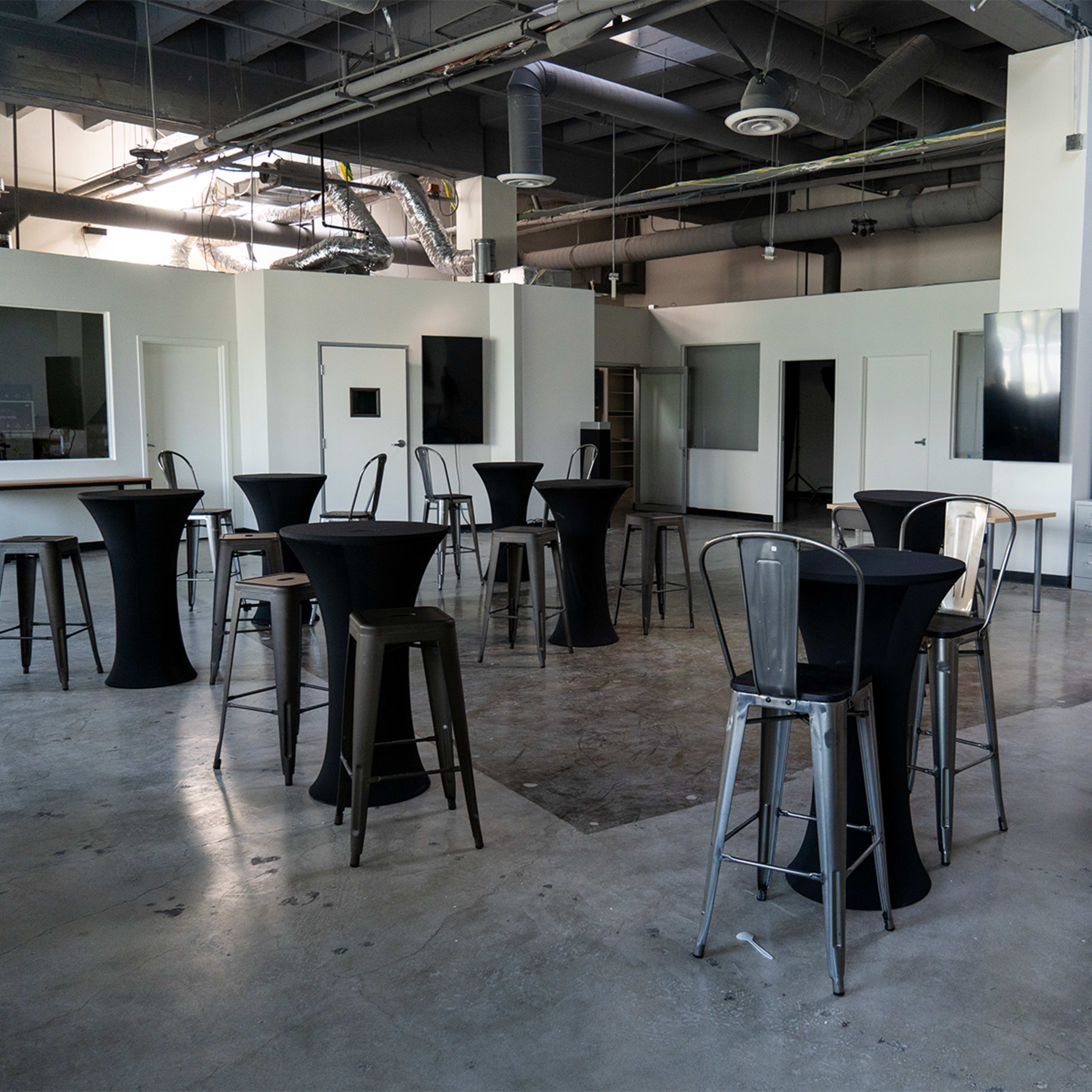 A spacious room with high tables and metal stools arranged in the center, surrounded by white walls and large windows.