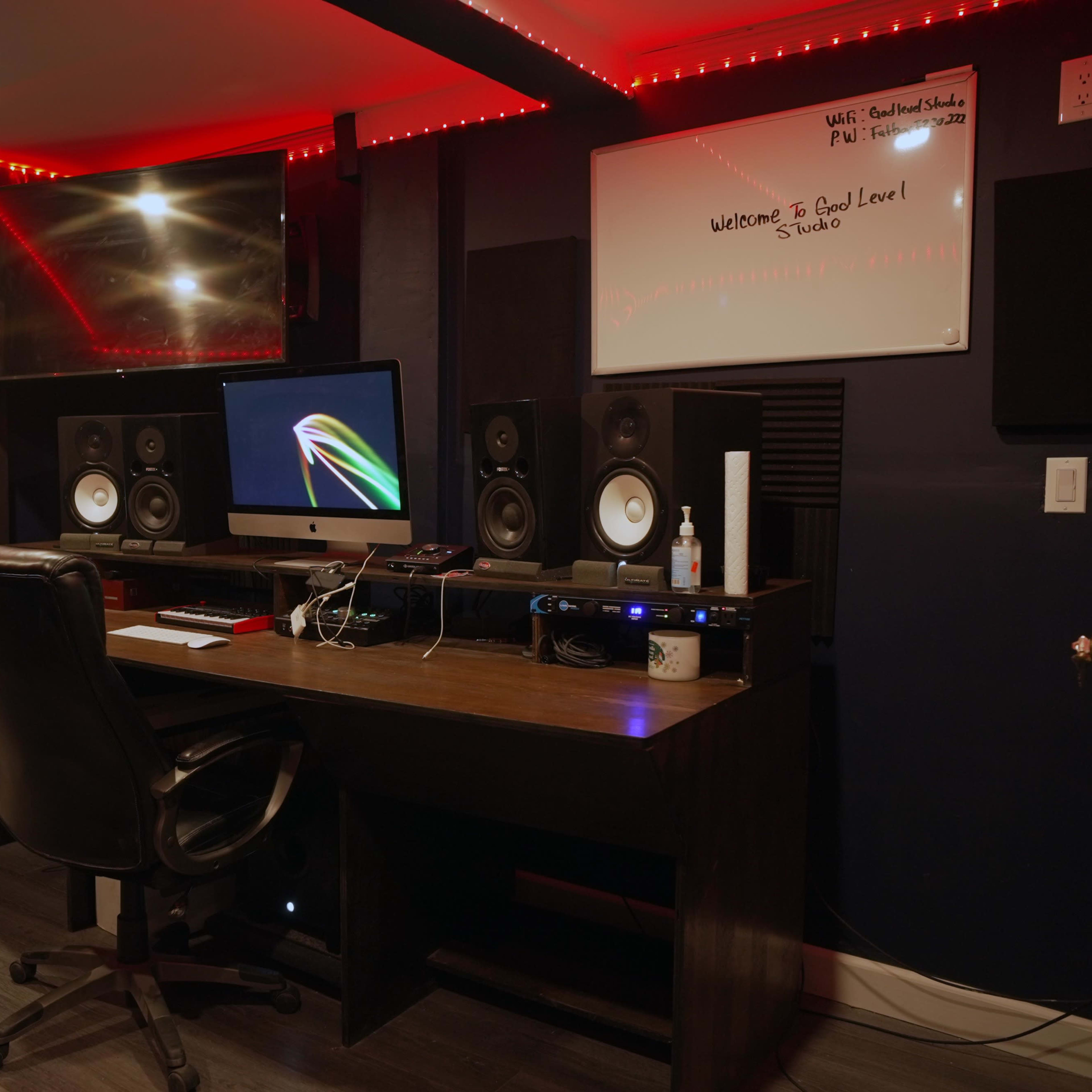 A dimly lit recording studio with a desk, a computer, and audio equipment, surrounded by soundproof panels and illuminated by red LED lights.