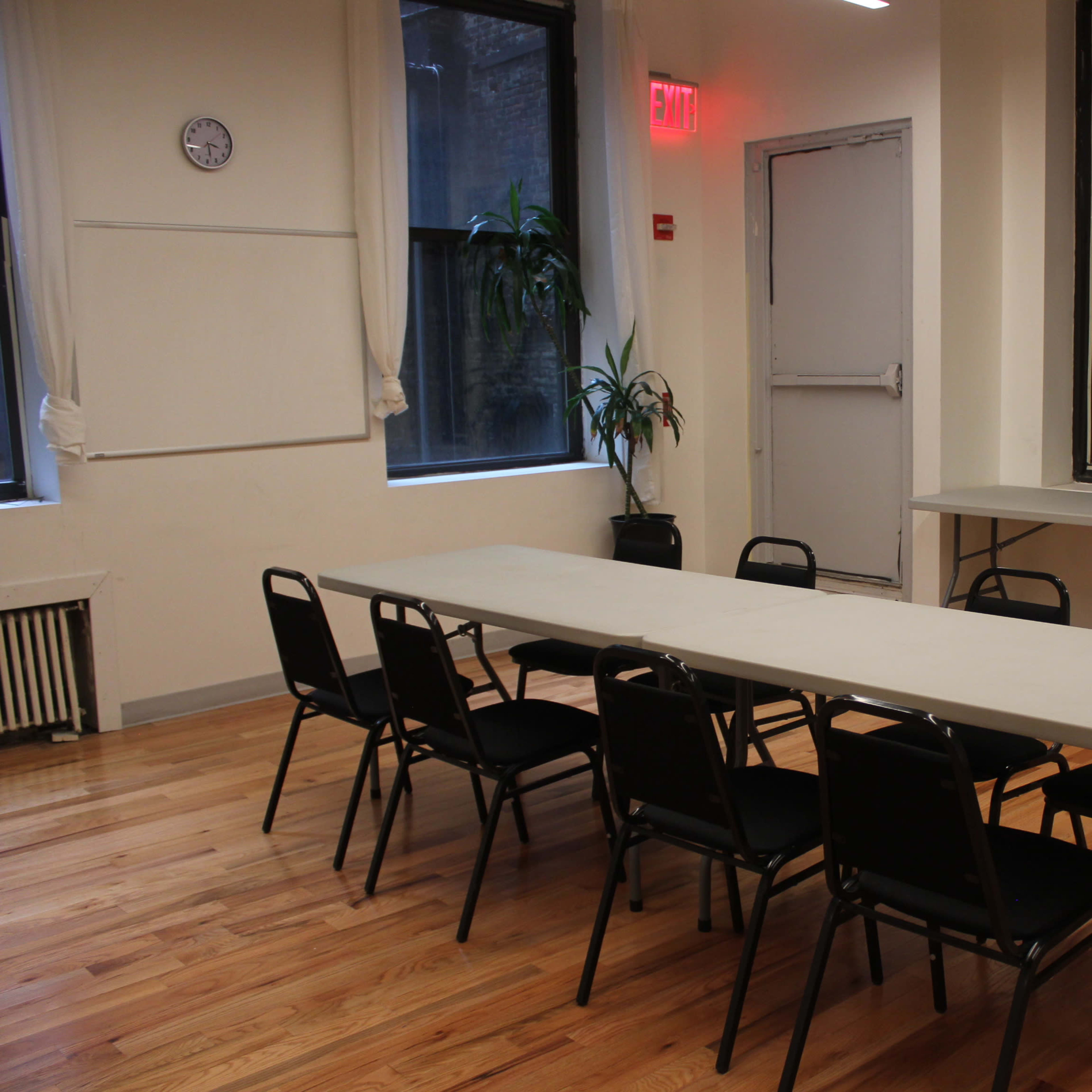 A large, empty table surrounded by black chairs is set up in a well-lit room with wooden floors and windows.