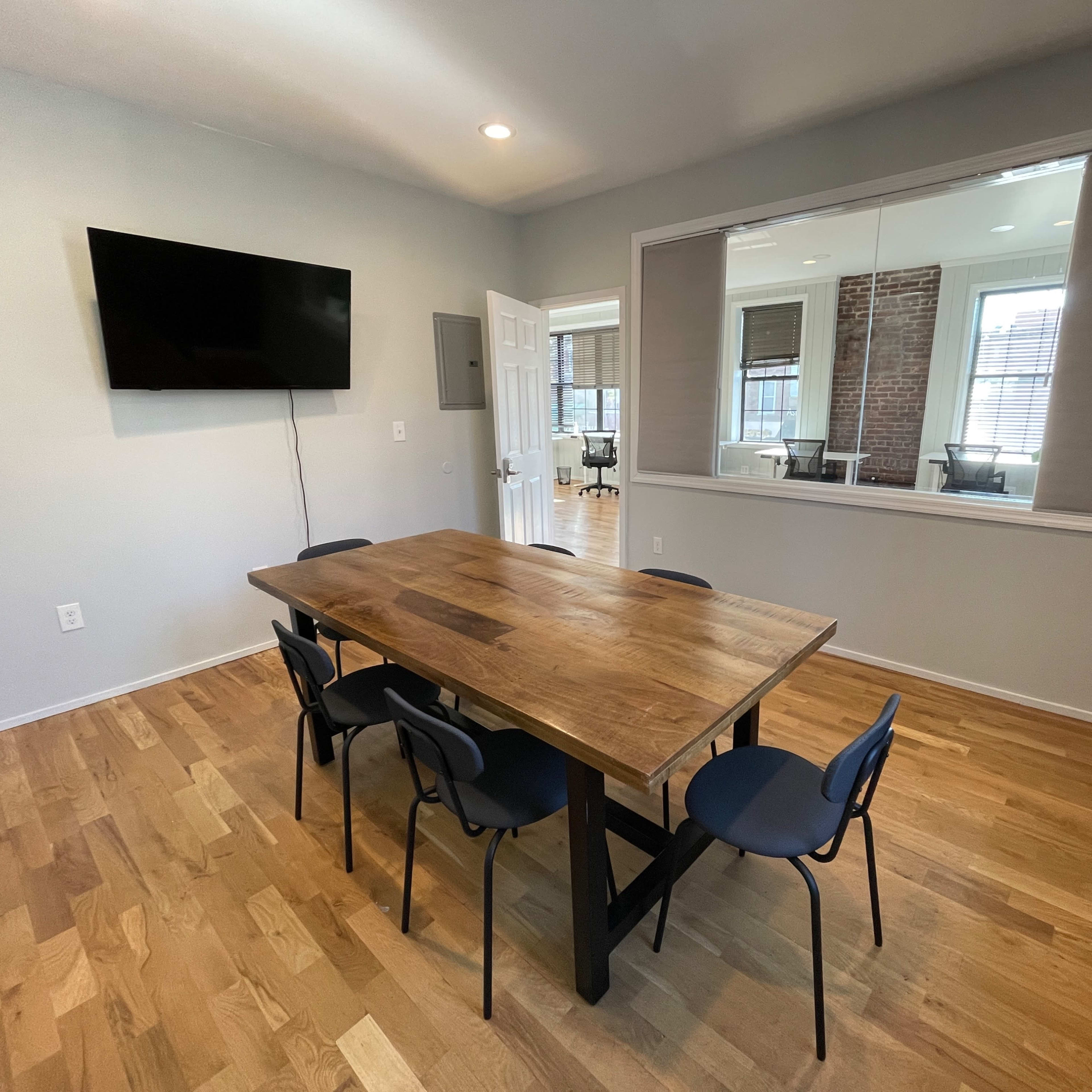 A simple, modern meeting room featuring a wooden table surrounded by chairs, with a television mounted on the wall and a large mirror reflecting part of the space.