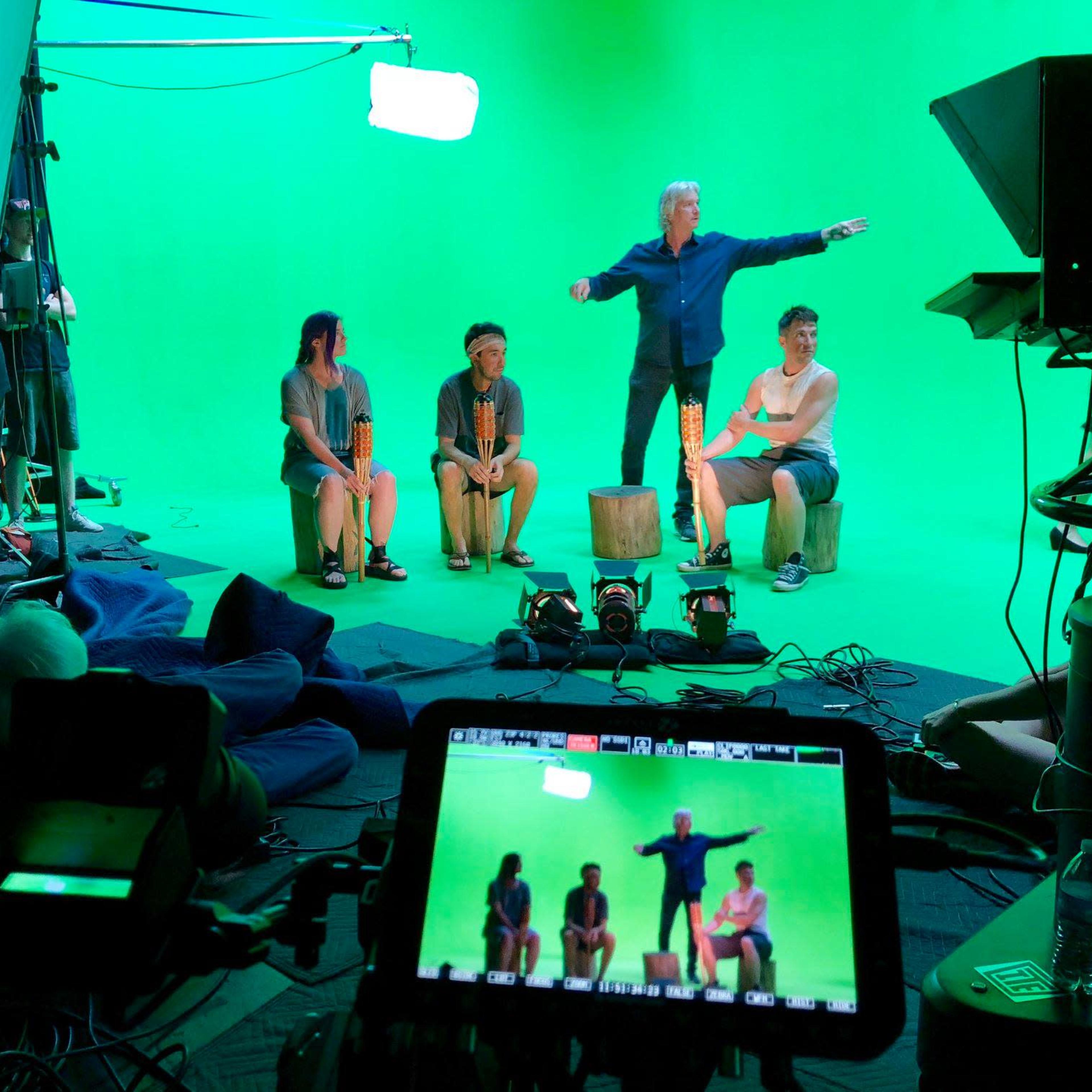 A group of individuals is positioned on a set with a green screen, while one person stands on a small elevated platform directing the scene.