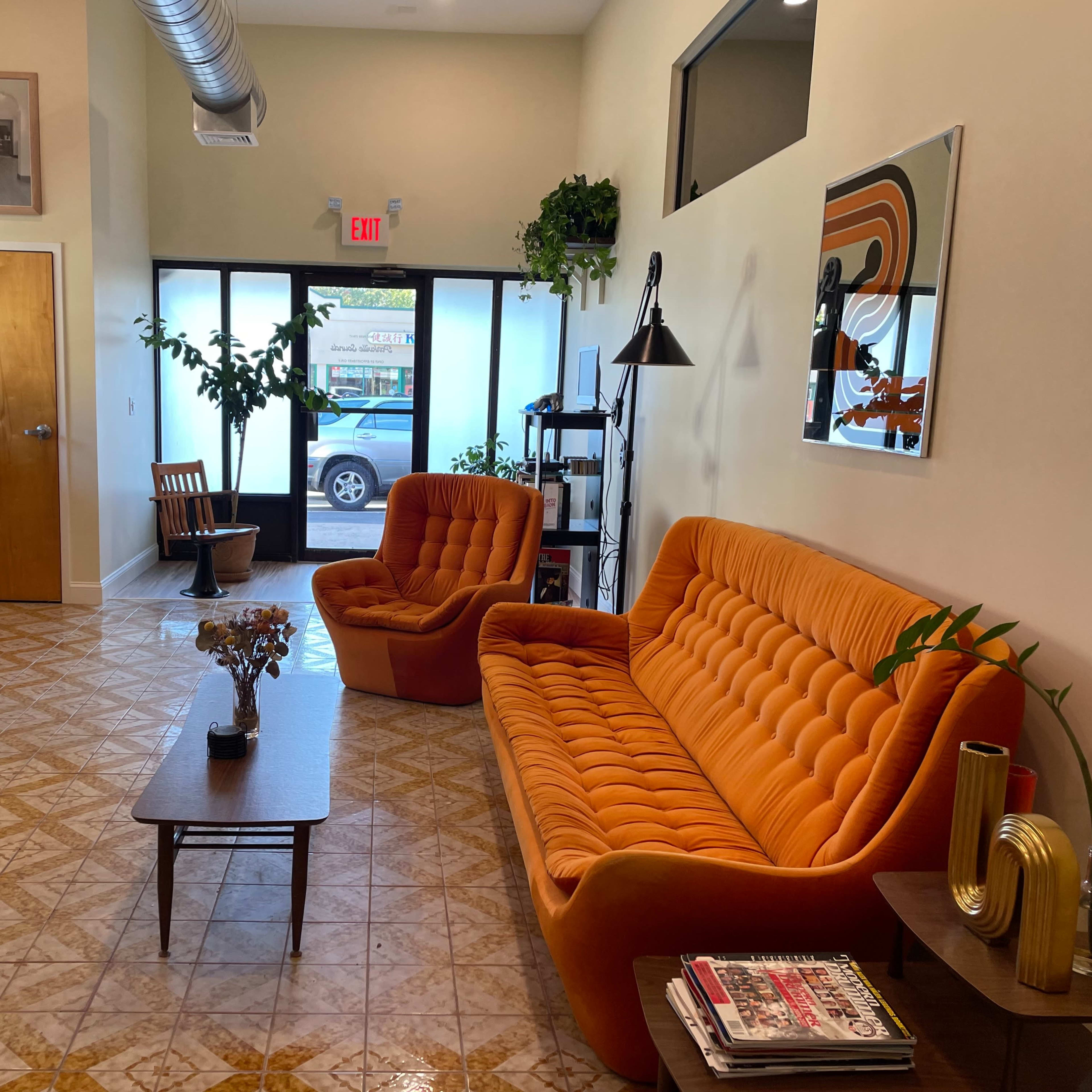 A bright, modern waiting area featuring an orange sofa, a wooden coffee table, and large windows with greenery visible outside.
