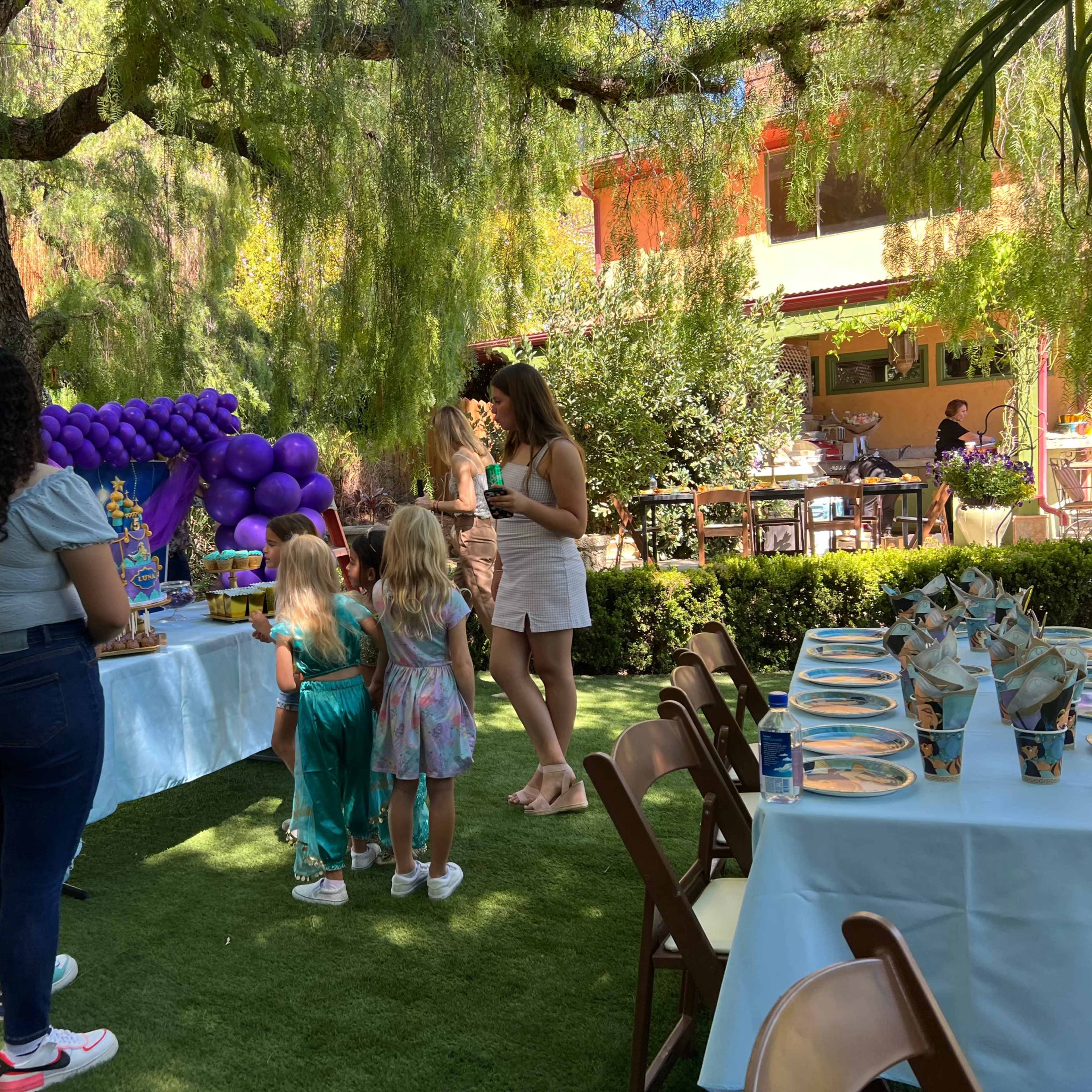 A family gathering is taking place in a garden, featuring a table with a decorated cake and colorful balloons, while two children look at the dessert setup.
