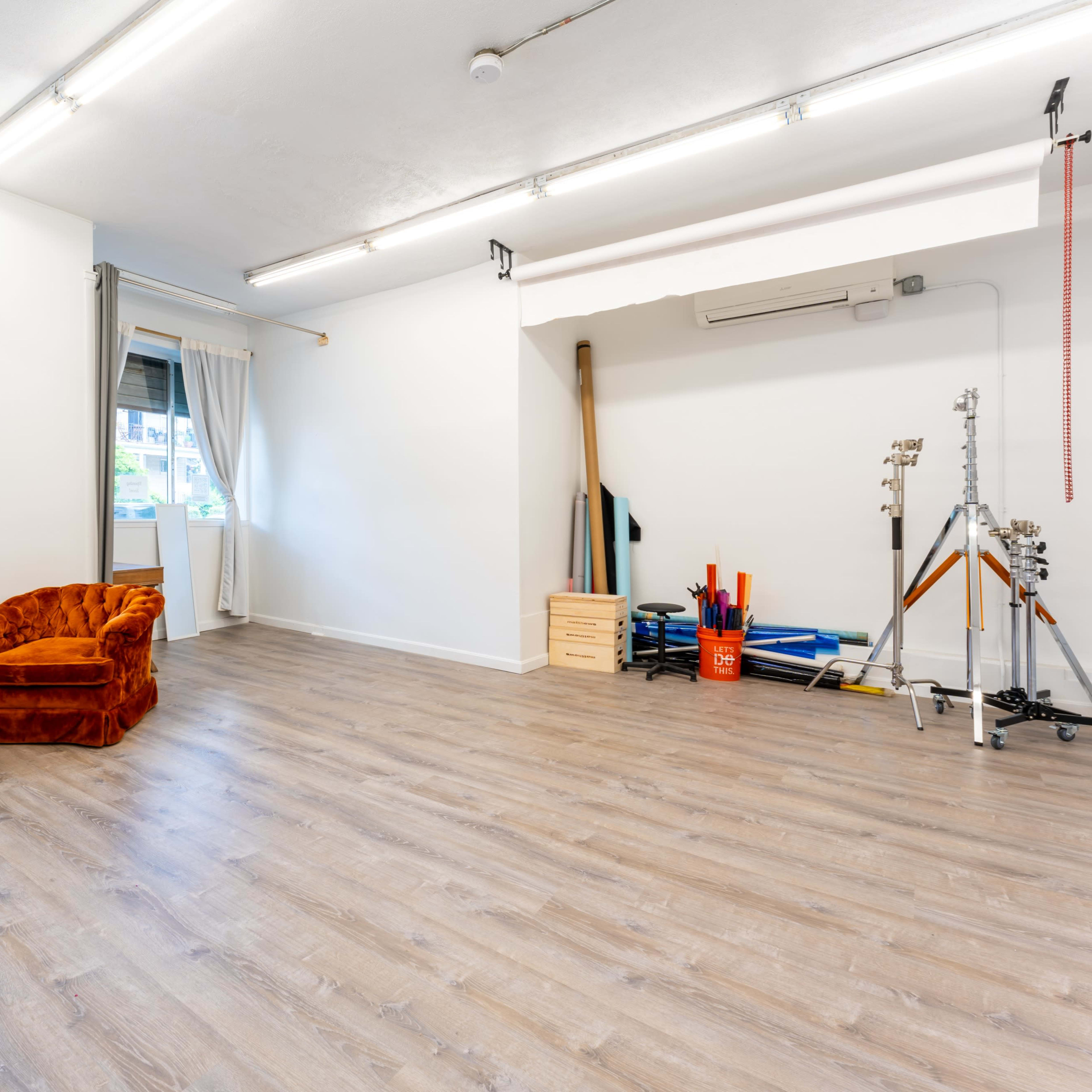 A bright, sparsely furnished room with wooden flooring, featuring a velvet orange chair, a window with curtains, and various exercise equipment in the corner.