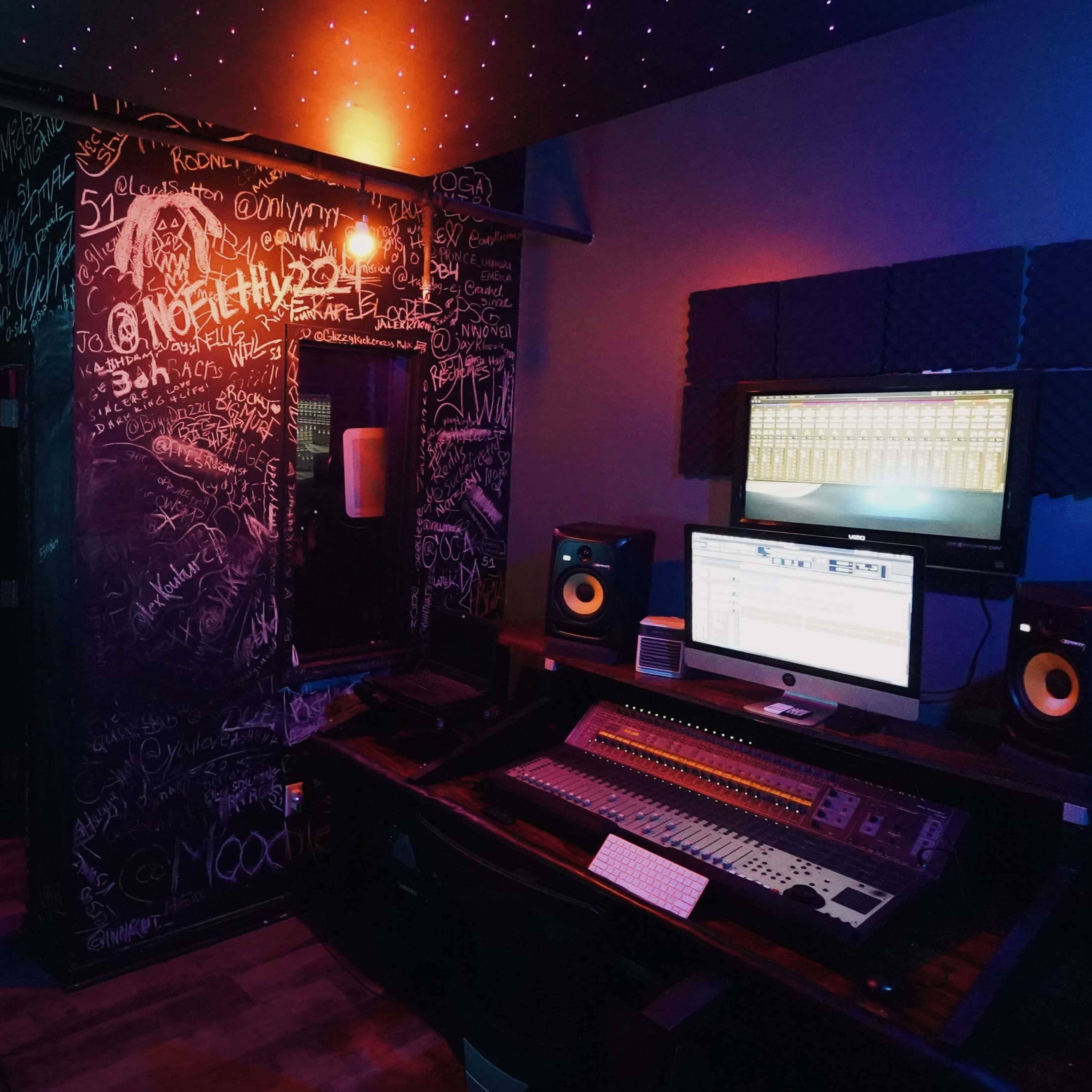 A music studio with a sound mixing desk, computer monitor displaying audio software, and a wall covered in chalkboard-style graffiti.