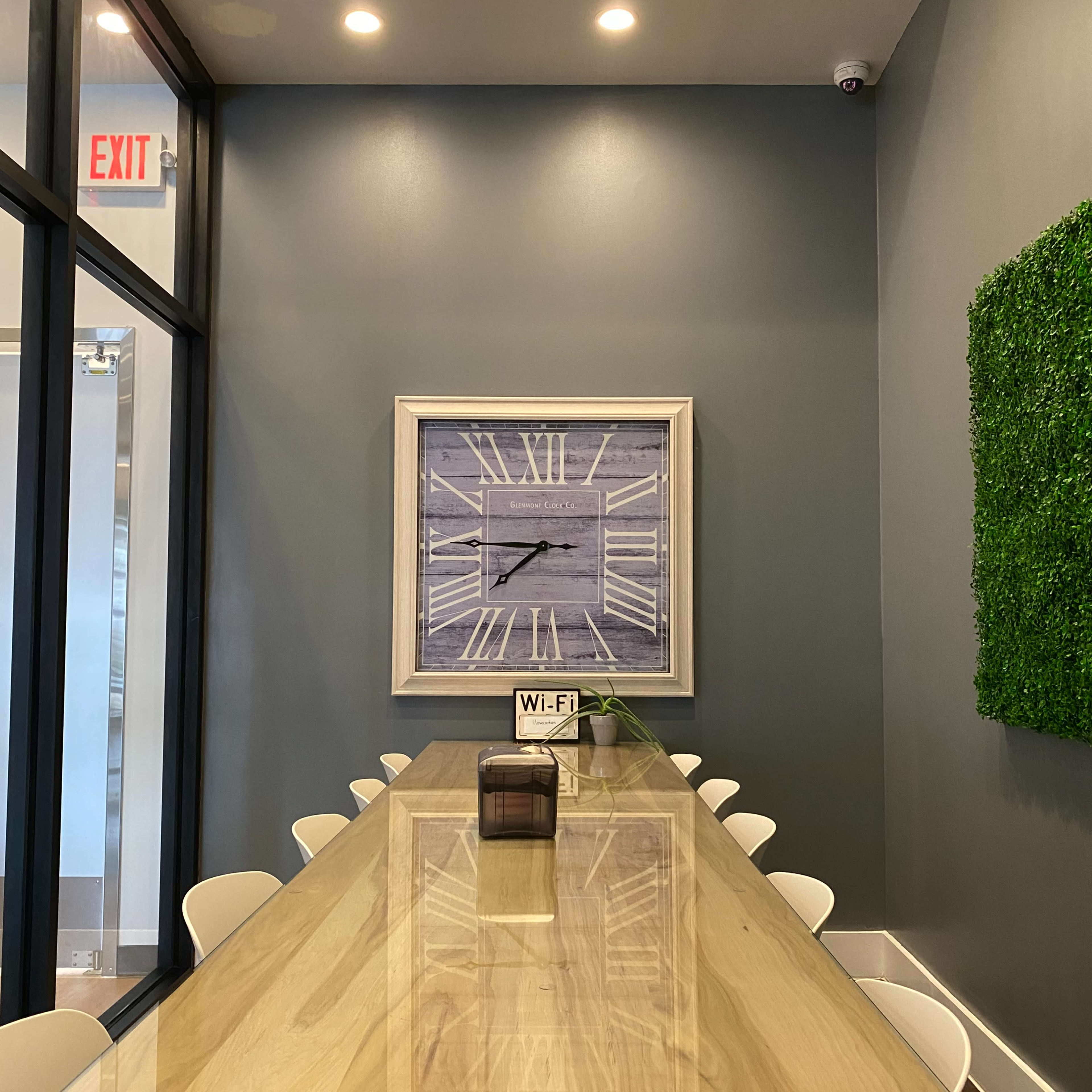 A long wooden table with white chairs is positioned in a well-lit room featuring a large wall clock and a green wall accent.