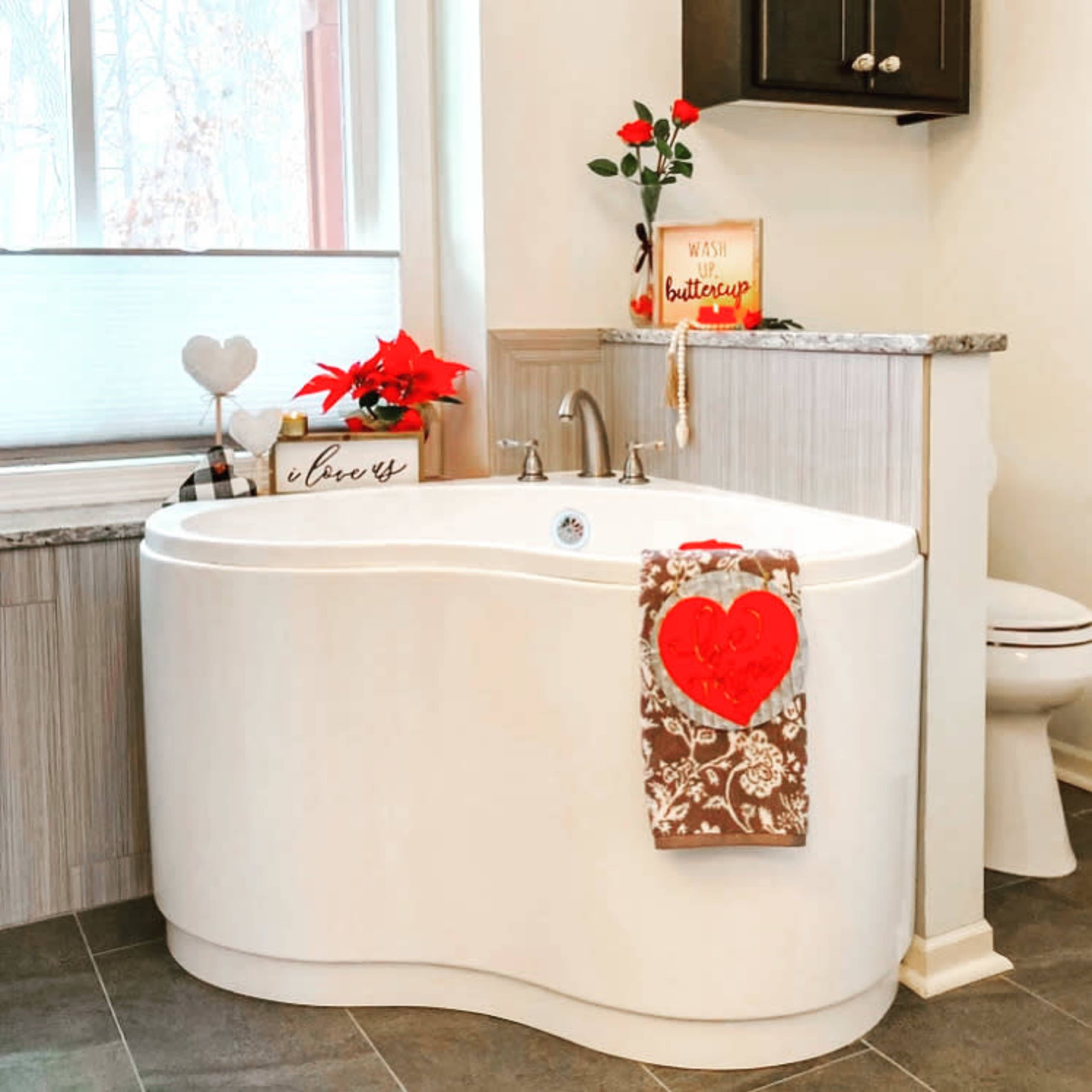 A modern bathroom featuring a white, curved bathtub with a red heart-shaped towel hanging from the side, and decorative items including a flower and a sign on the counter.