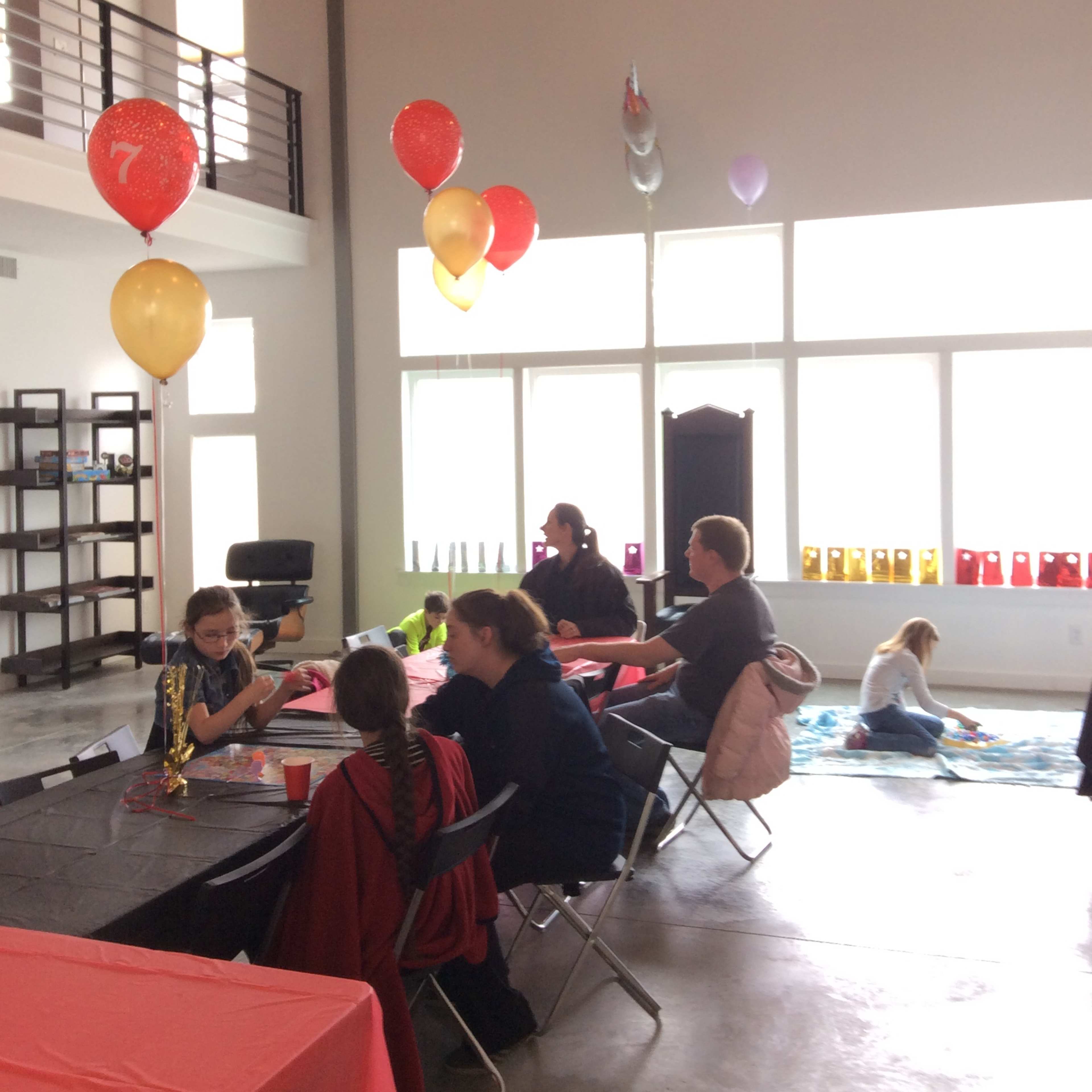 A group of people sits at tables in a spacious room decorated with colorful balloons for a celebration.