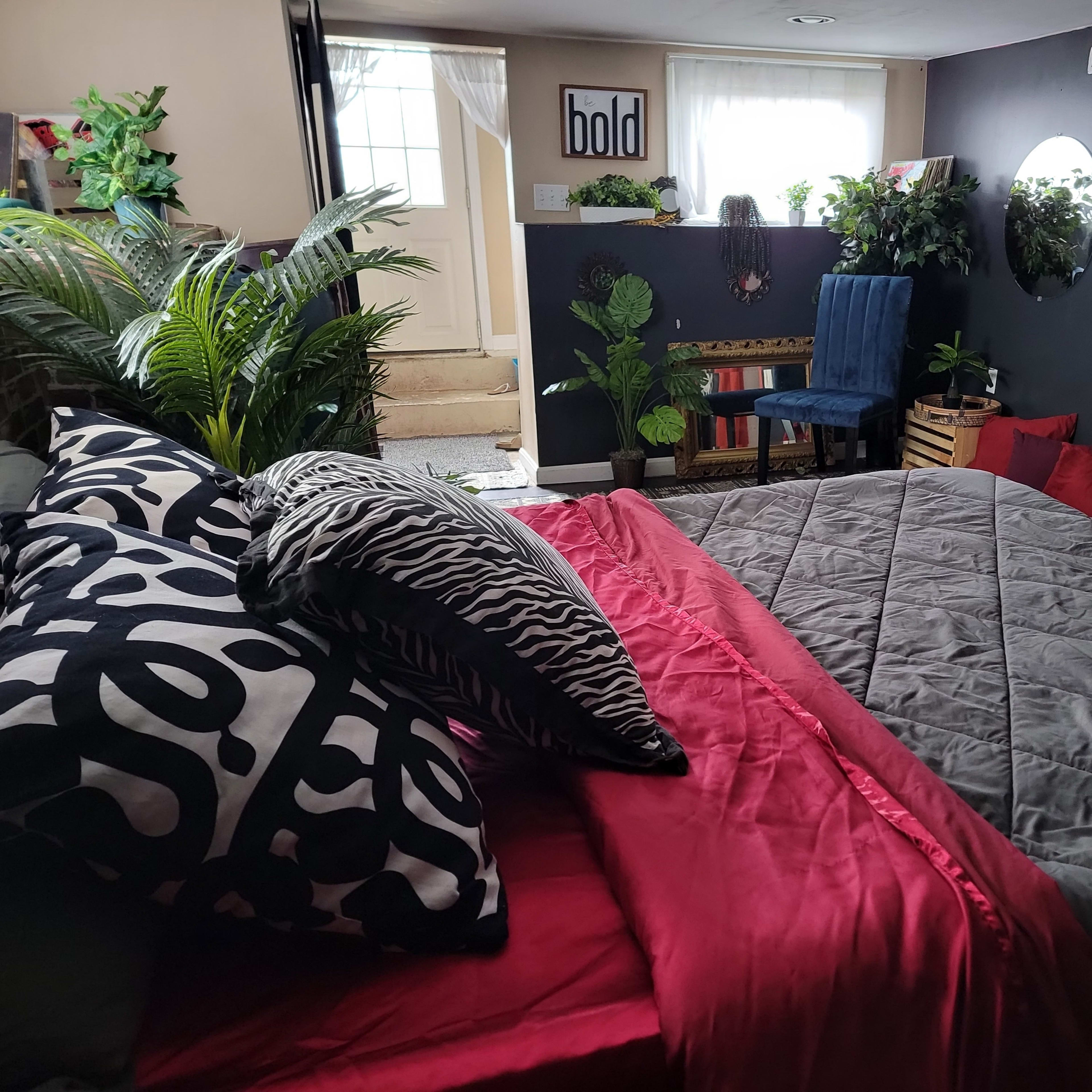 A cozy bedroom with a large bed covered in red and gray bedding, a decorative wall with greenery, and a seating area featuring a blue chair and various plants.