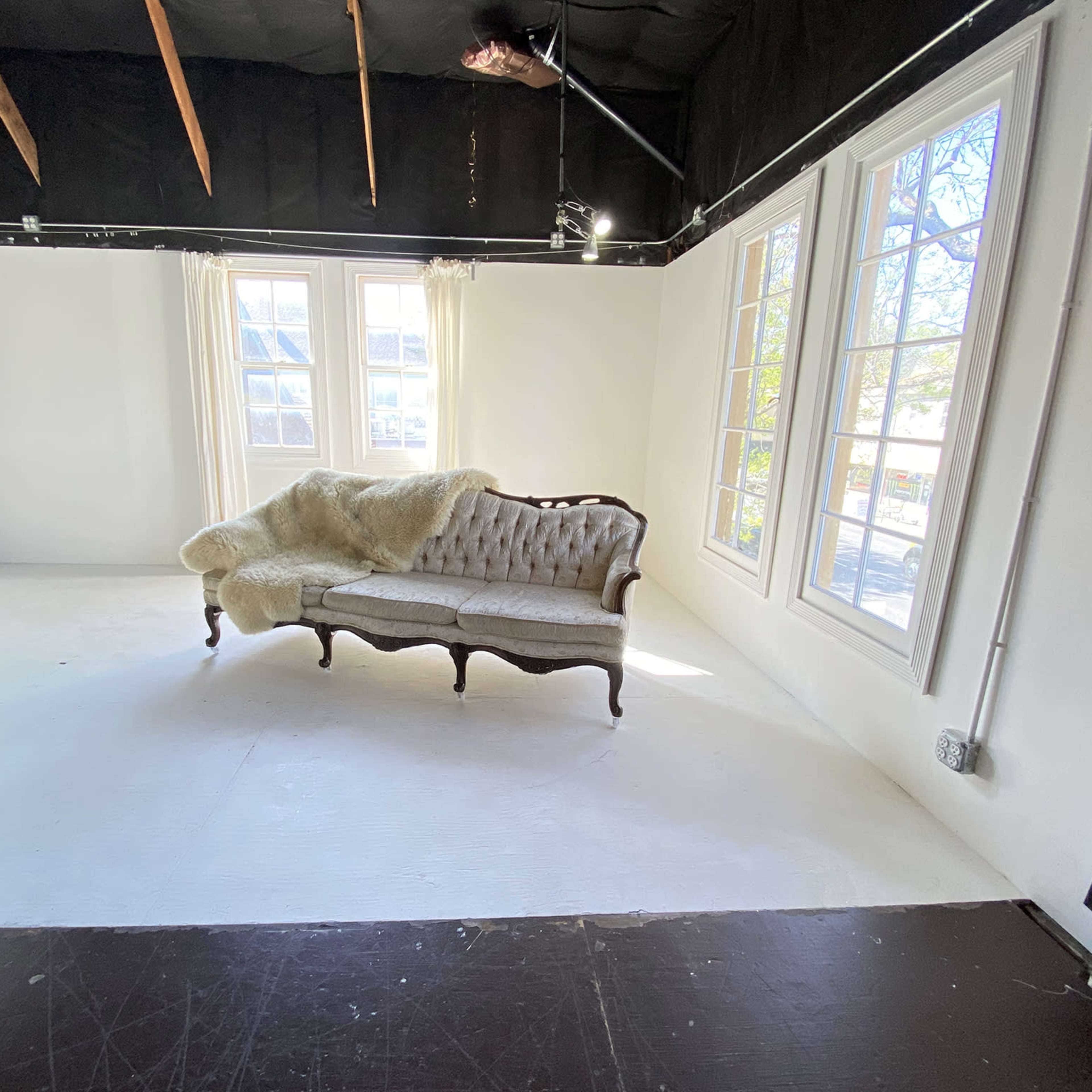 A vintage tufted sofa with a faux fur throw is positioned in an empty, well-lit room with large windows.