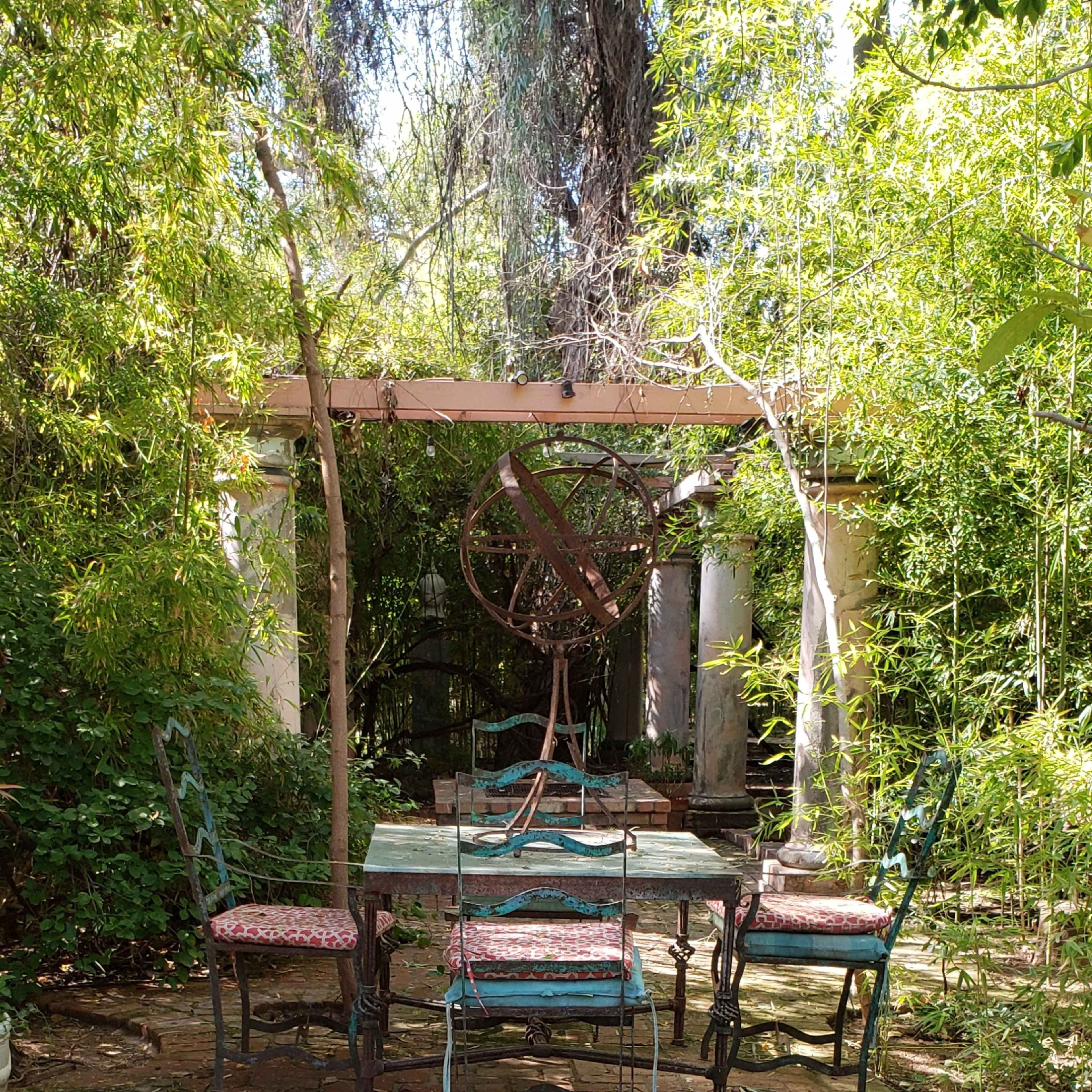 A secluded garden patio with a stone pathway, surrounded by lush greenery and featuring a table with chairs under a wooden pergola.