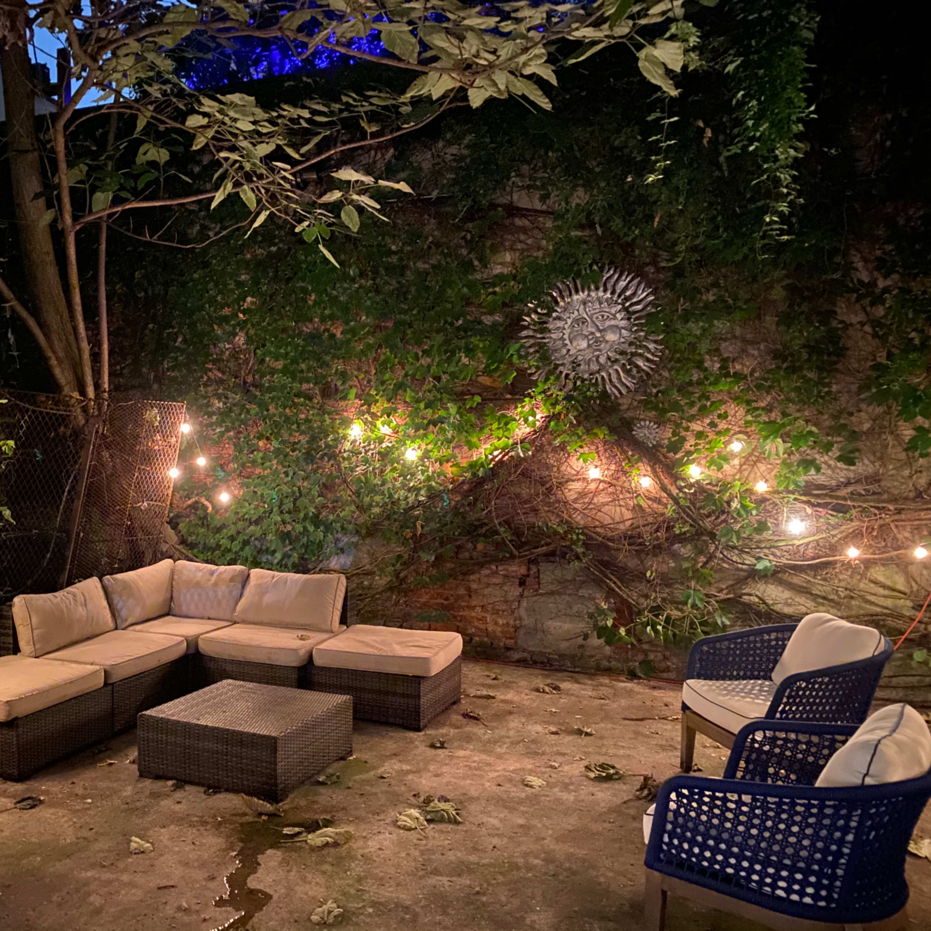 A garden patio area with a sectional sofa, coffee table, and several chairs surrounded by greenery and string lights.
