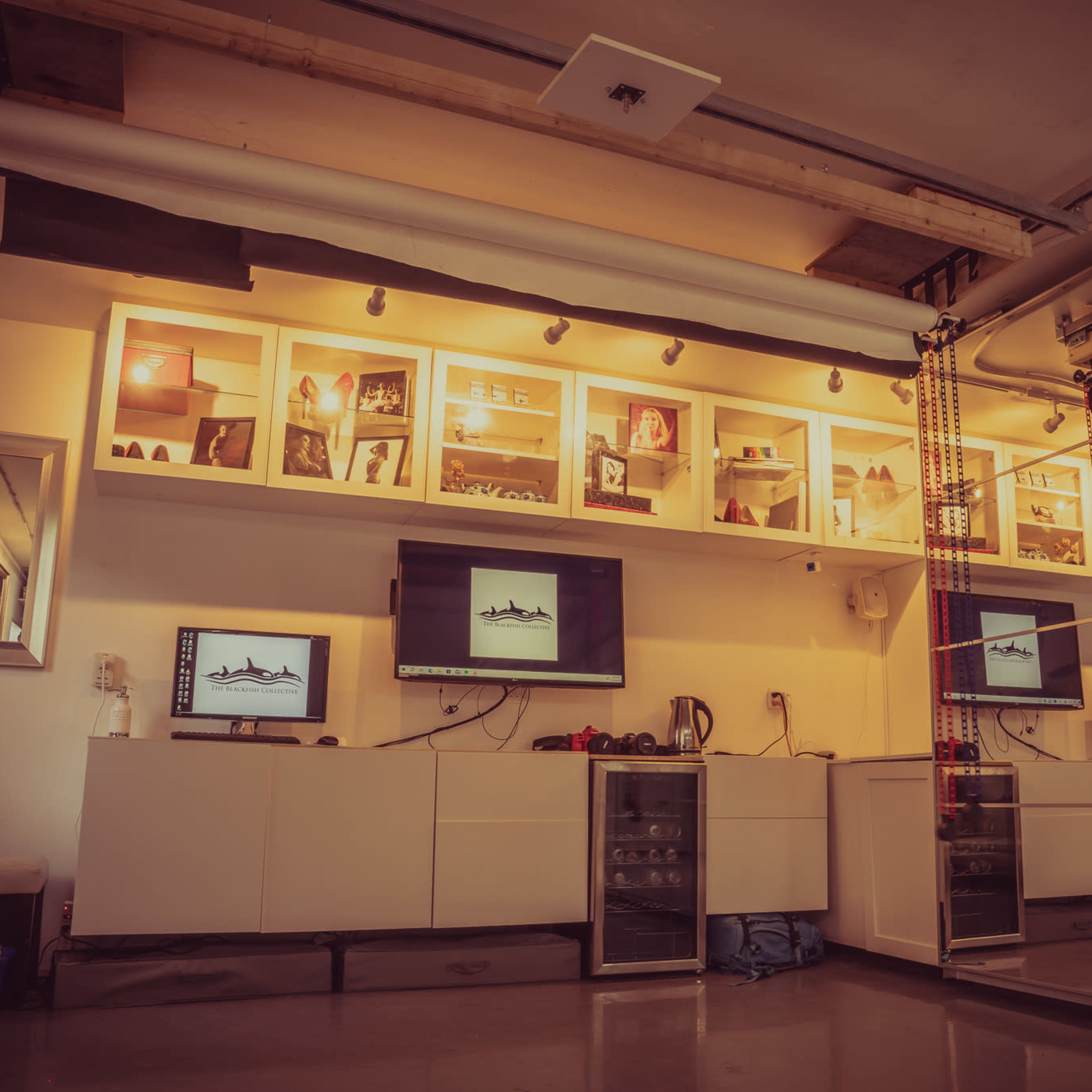 A modern room with a wall-mounted shelving unit, two flat-screen monitors, and a small refrigerator beneath them.